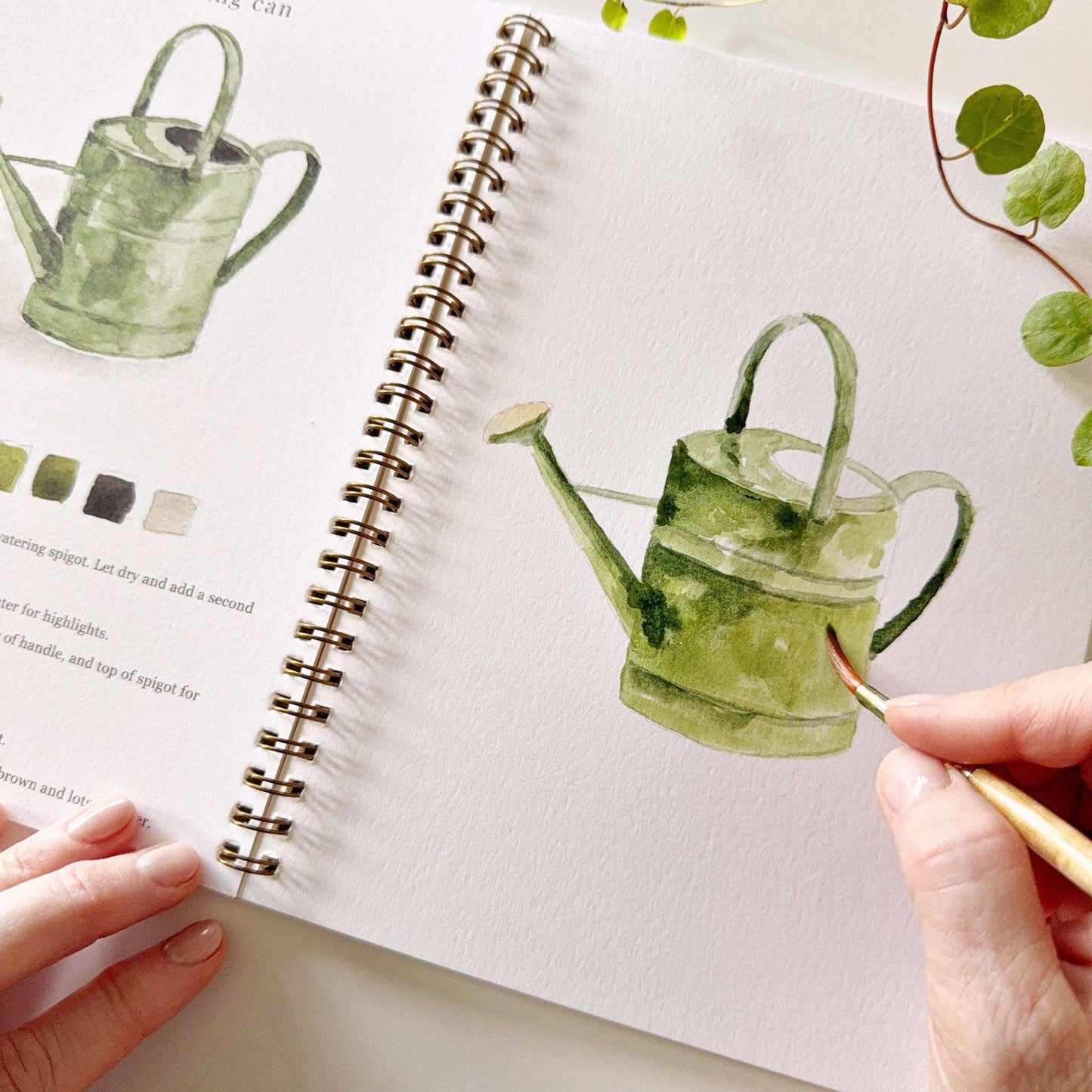 A garden watercolor workbook with step-by-step garden sketches, here with a close-up on the watering can page, with someone painting with a brush