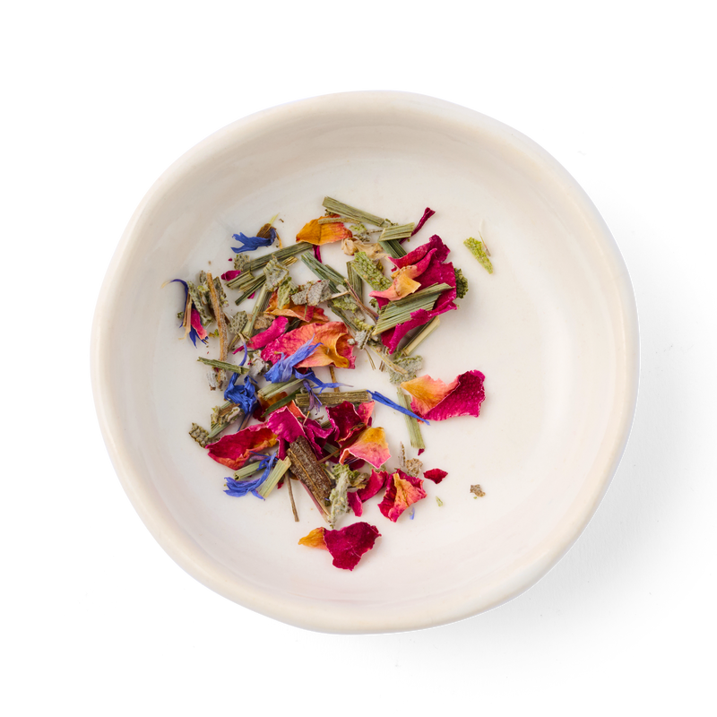White bowl with colorful dried flowers and plants on a white background. The plants are sage, lemongrass, rose petals and blue cornflower, which are the ingredients of the Honey Honey herbal tea from the brand Dona.