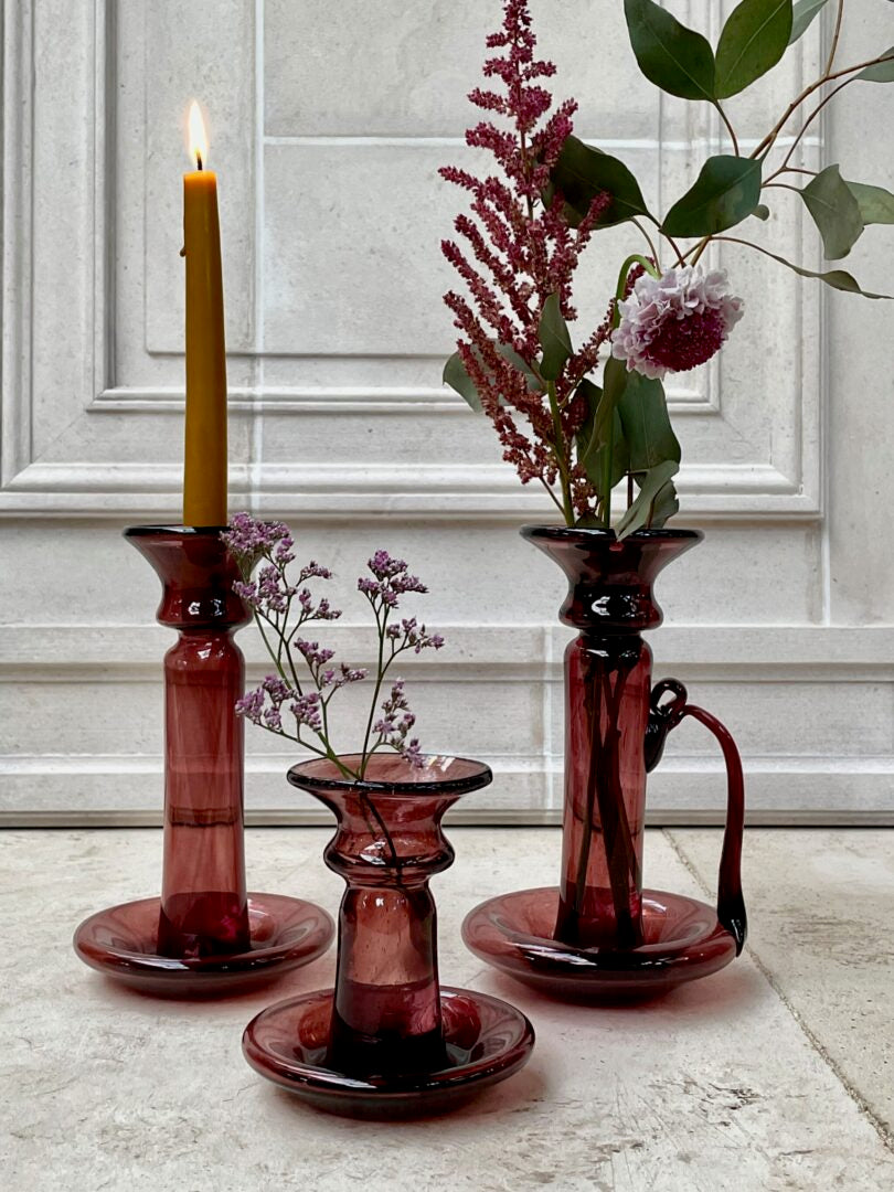 Set of three raspberry pink mouth-blown glass candle holders with flowers and a candle on a light-colored surface.
The candle holder on the left is with a handle.