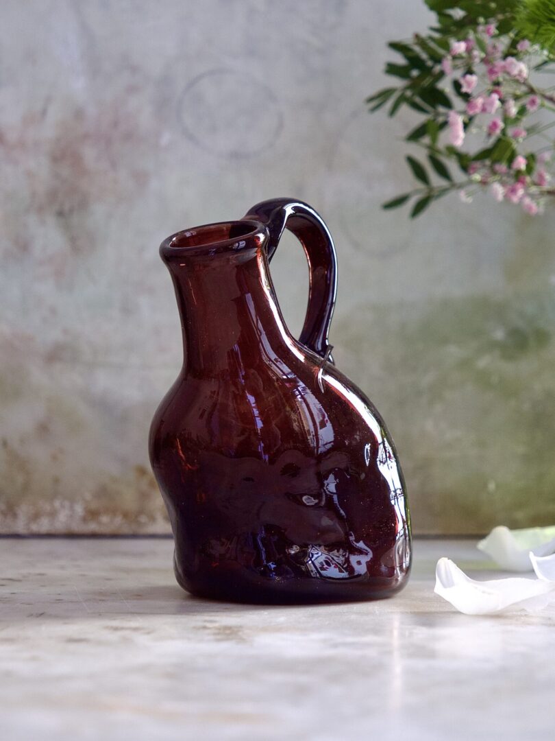 Brown mouth-blown glass pitcher with a tilt on a marble surface with a blurred floral background.