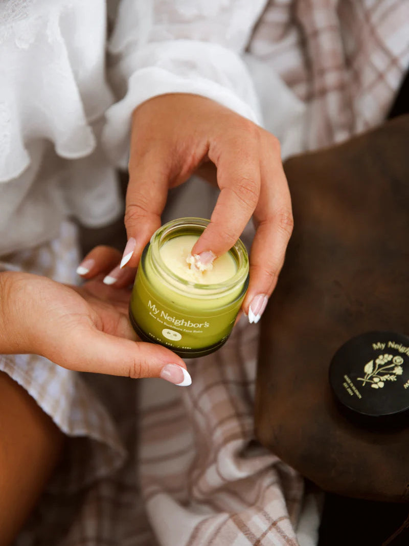 Person holding a green jar of balm labeled 'My Neighbor's' with a blurred background