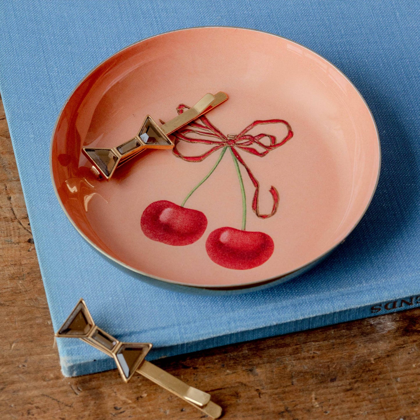 A round peach pink enamel tray with a cherry design in the center, depicted with a red bow and two cherry fruits, holding hair accessories or jewellery, on a blue surface and a wooden table.