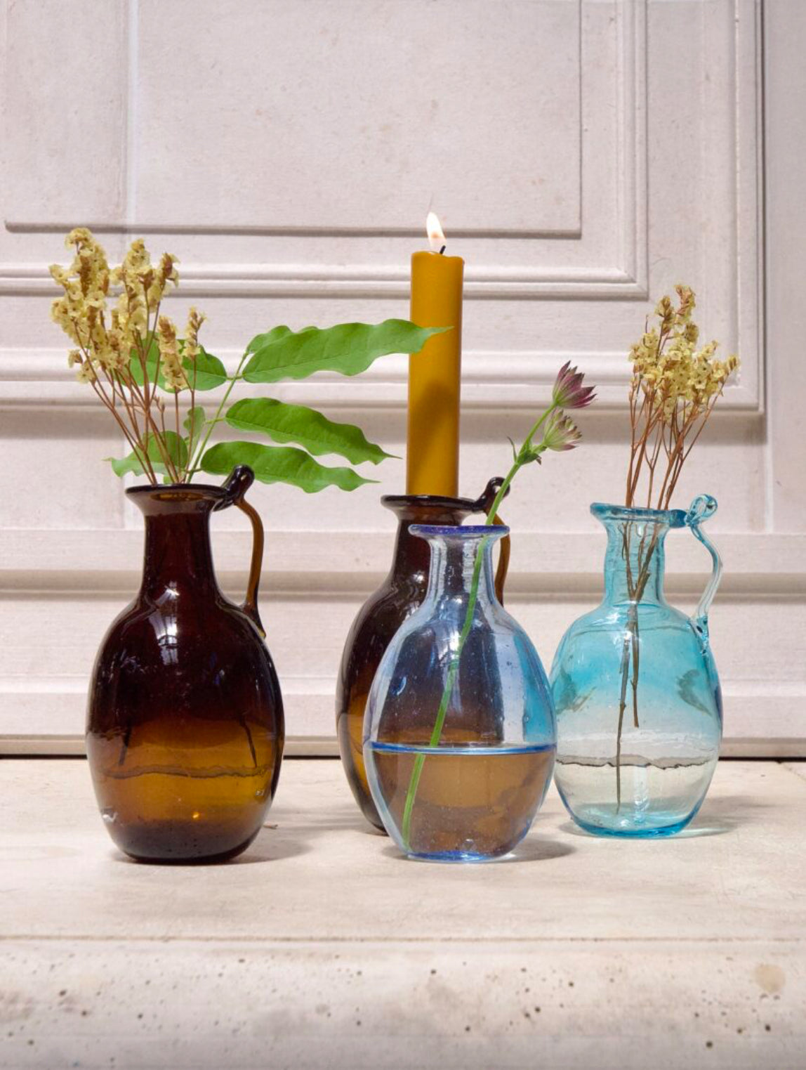 Four glass vases with plants and a candle on a light surface.