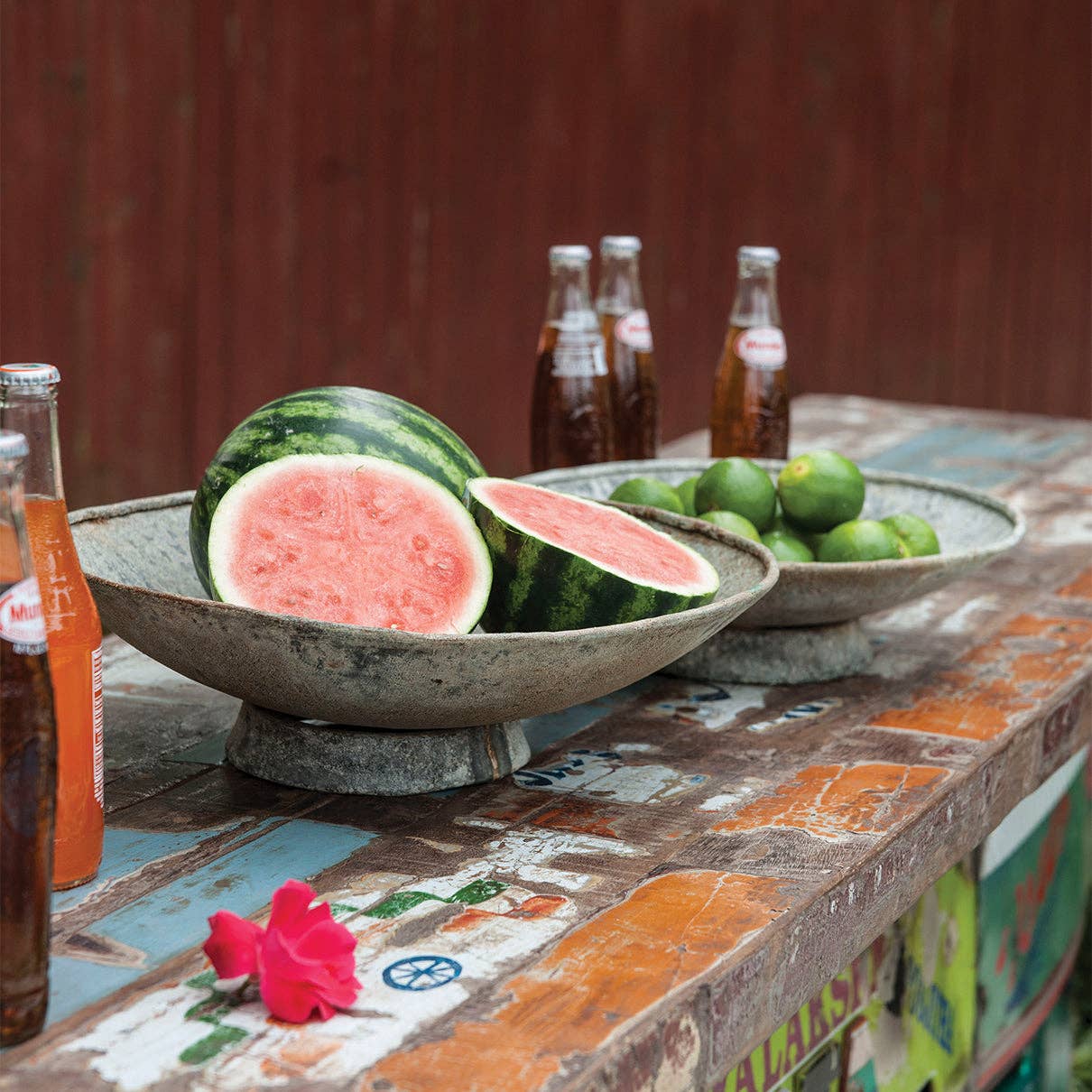 Two antique-style metal saucer planters with a rustic finish, one containing watermelons and one containing limes, on a colorful countertop with soda bottles nearby.
