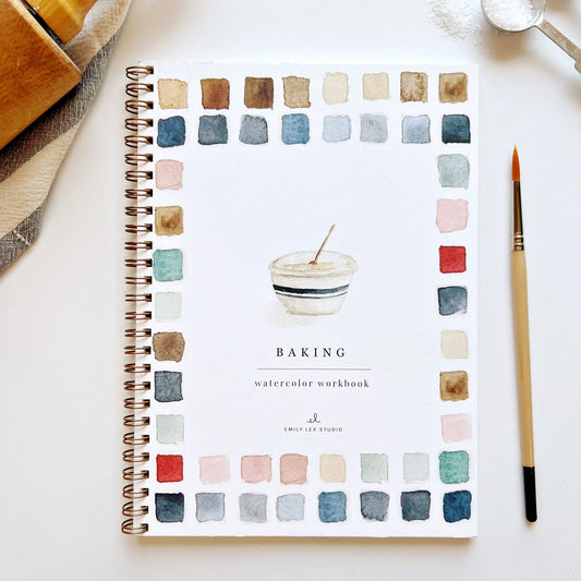 A watercolor workbook titled 'BAKING' with various colored watercolor swatches on the cover, along with a paintbrush and a spiral binding visible, placed on a white surface.