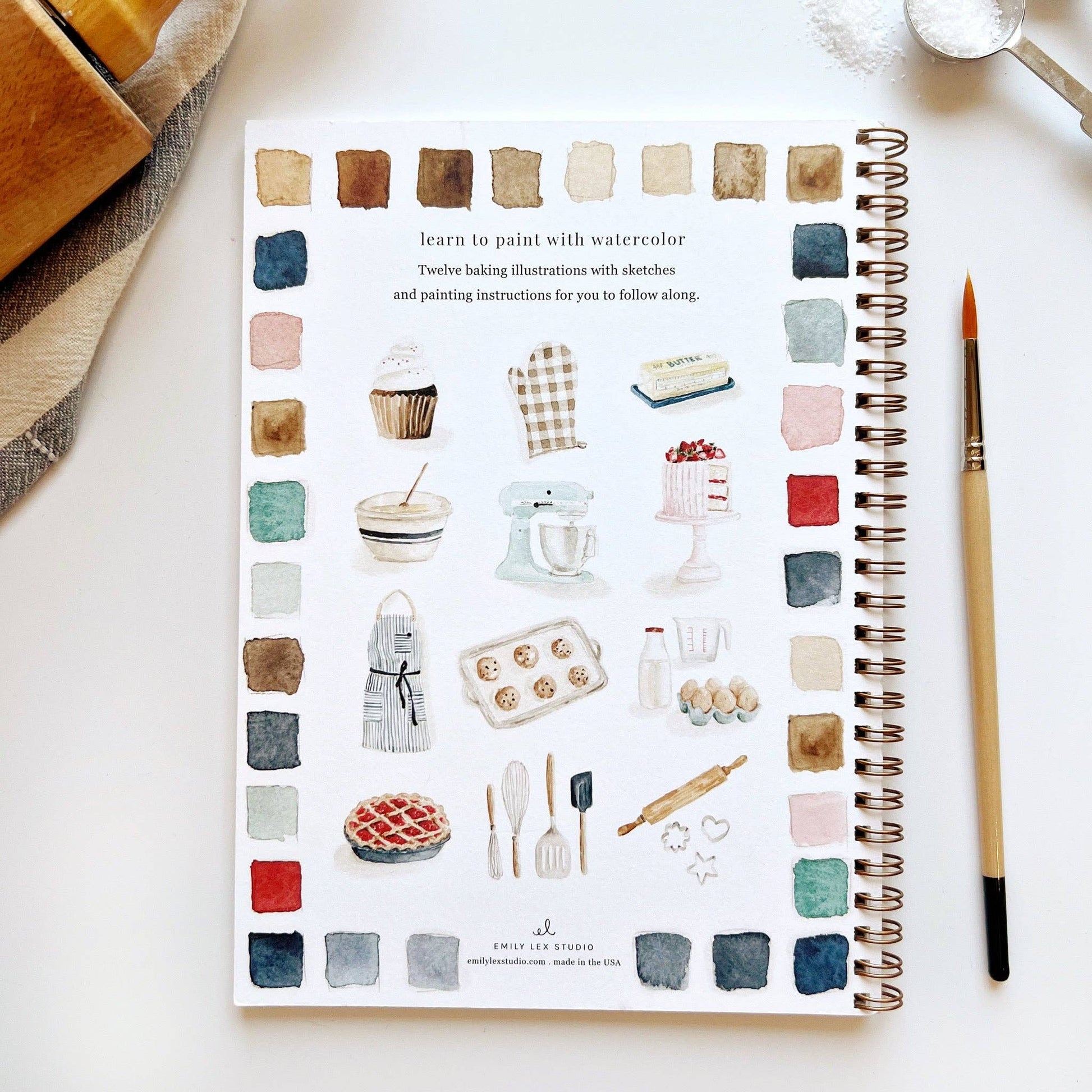 Baking Watercolor Workbook with baking-themed illustrations, color swatches and spiral binding. A watercolor brush is lying next to the book.
