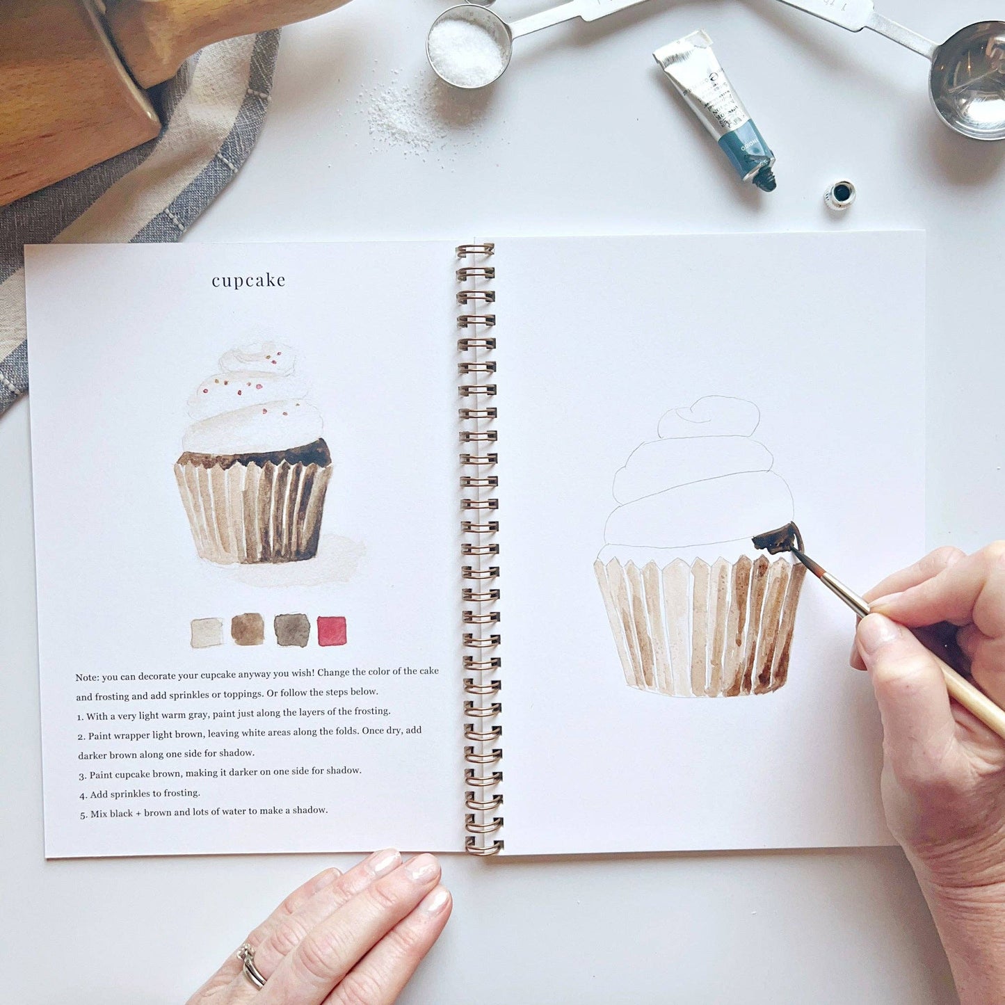 Creative watercolor activity book with step-by-step baking-themed sketches, here on the cupcake page, with someone painting with a brush
