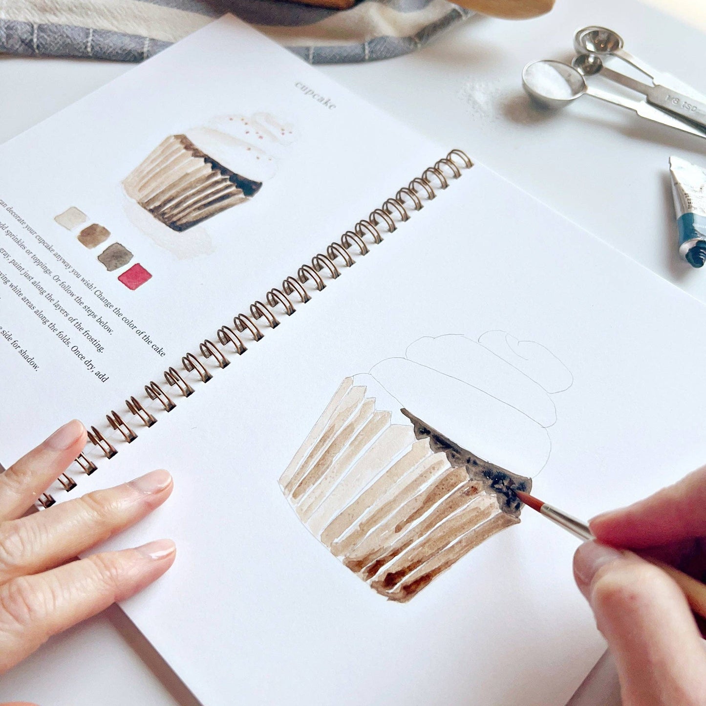 A baking watercolor workbook with step-by-step baking-themed sketches, here with a close-up on the cupcake page, with someone painting with a brush