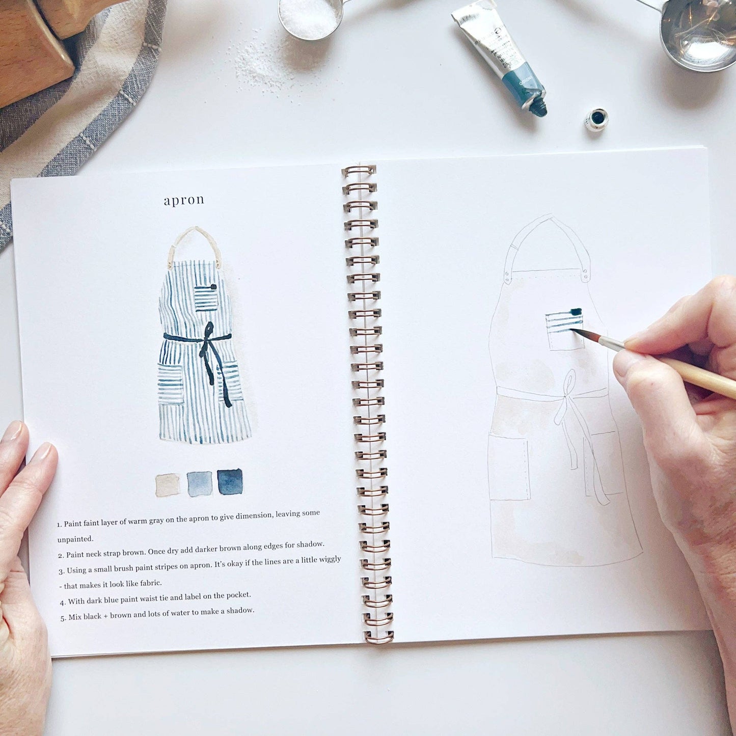 A baking watercolor workbook with step-by-step baking-themed sketches, here on the apron page