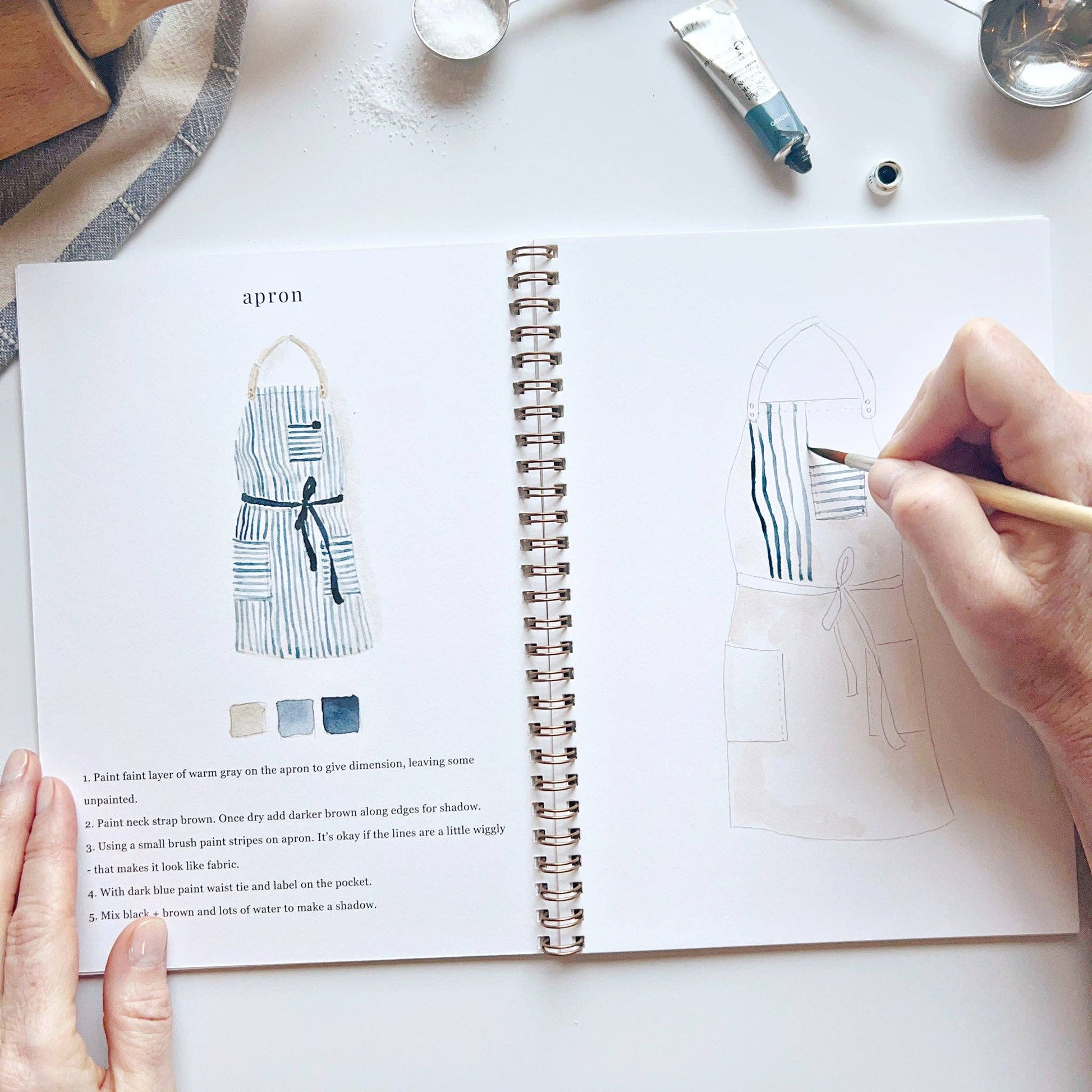 A baking watercolor workbook with step-by-step baking-themed sketches, here on the apron page, with someone painting with a brush.