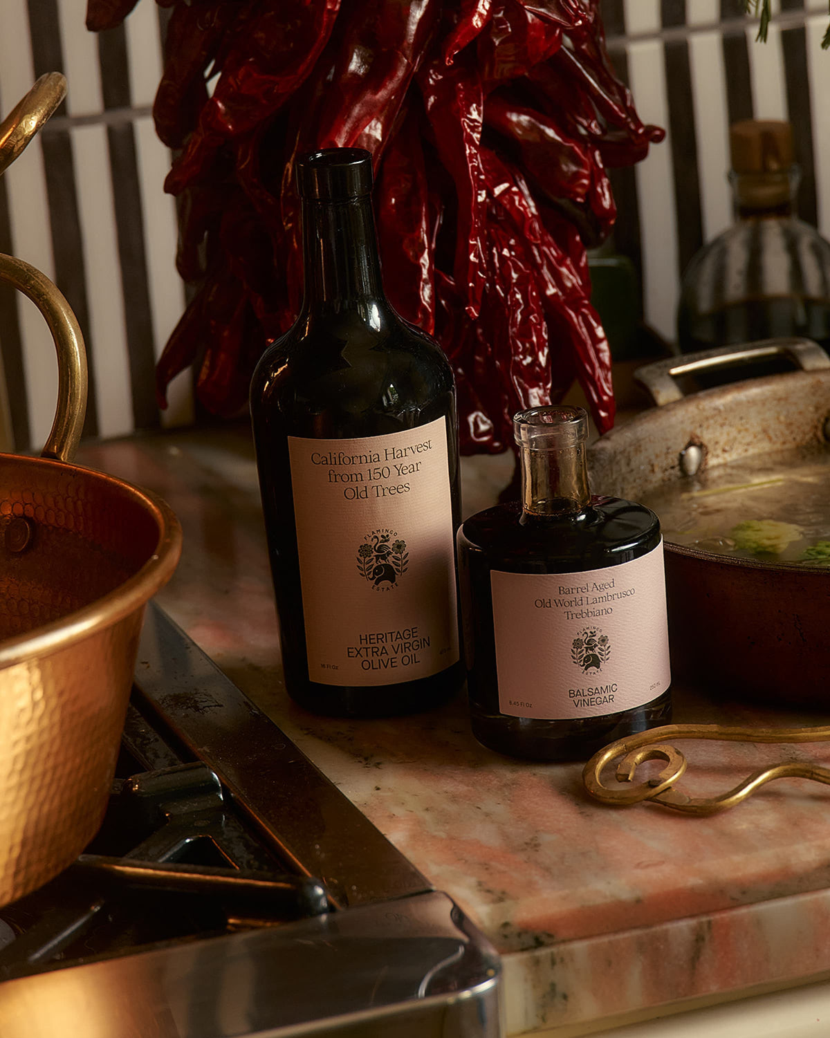 A bottle of balsamic vinegar next to a bottle of olive oil from Flamingo Estate  on a rustic surface with copper pots and red chili peppers in the background.