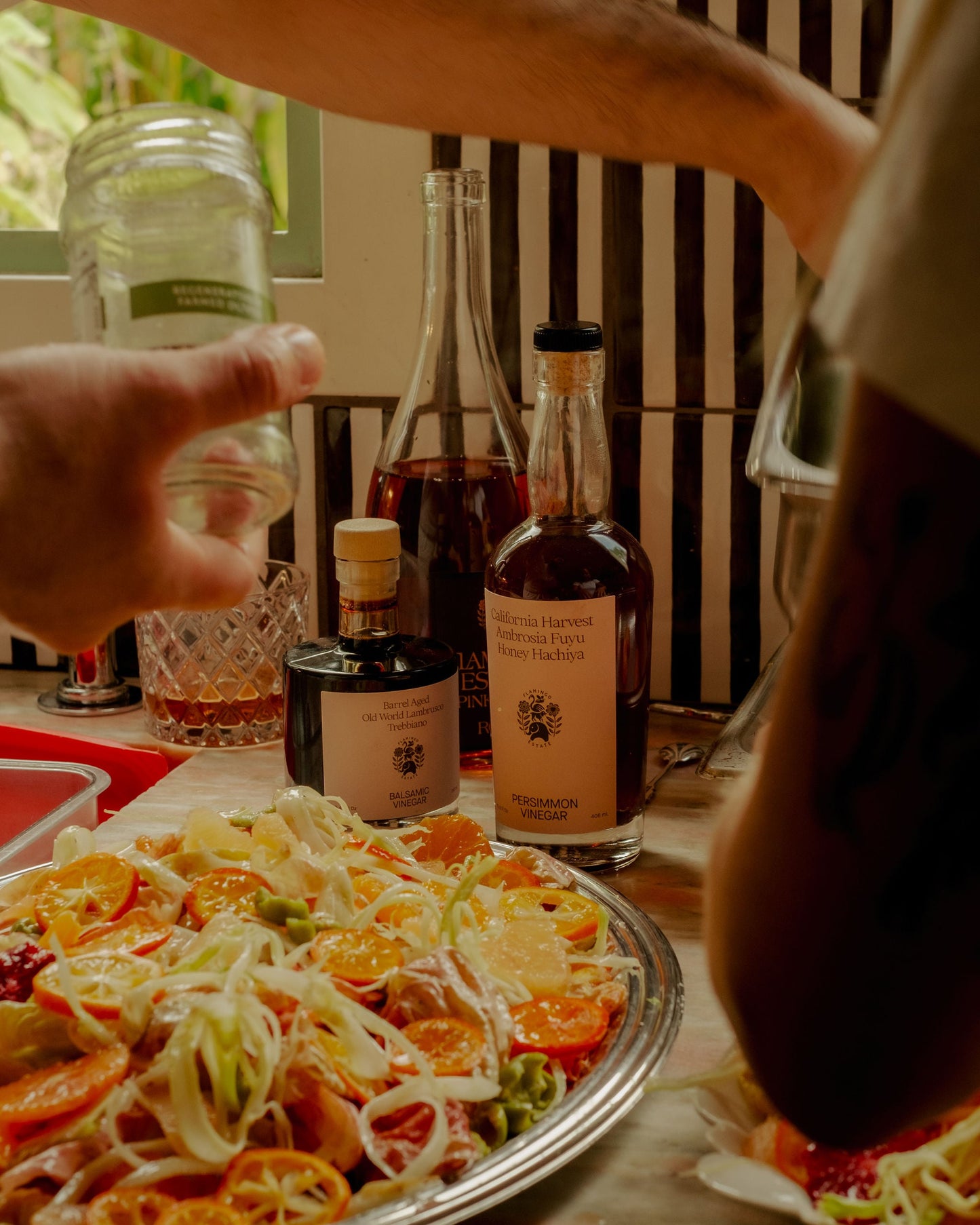 Table with a salad, bottles of balsamic vinegar and persimmon fruit vinegar from Flamingo Estate, and a hand holding a glass jar.