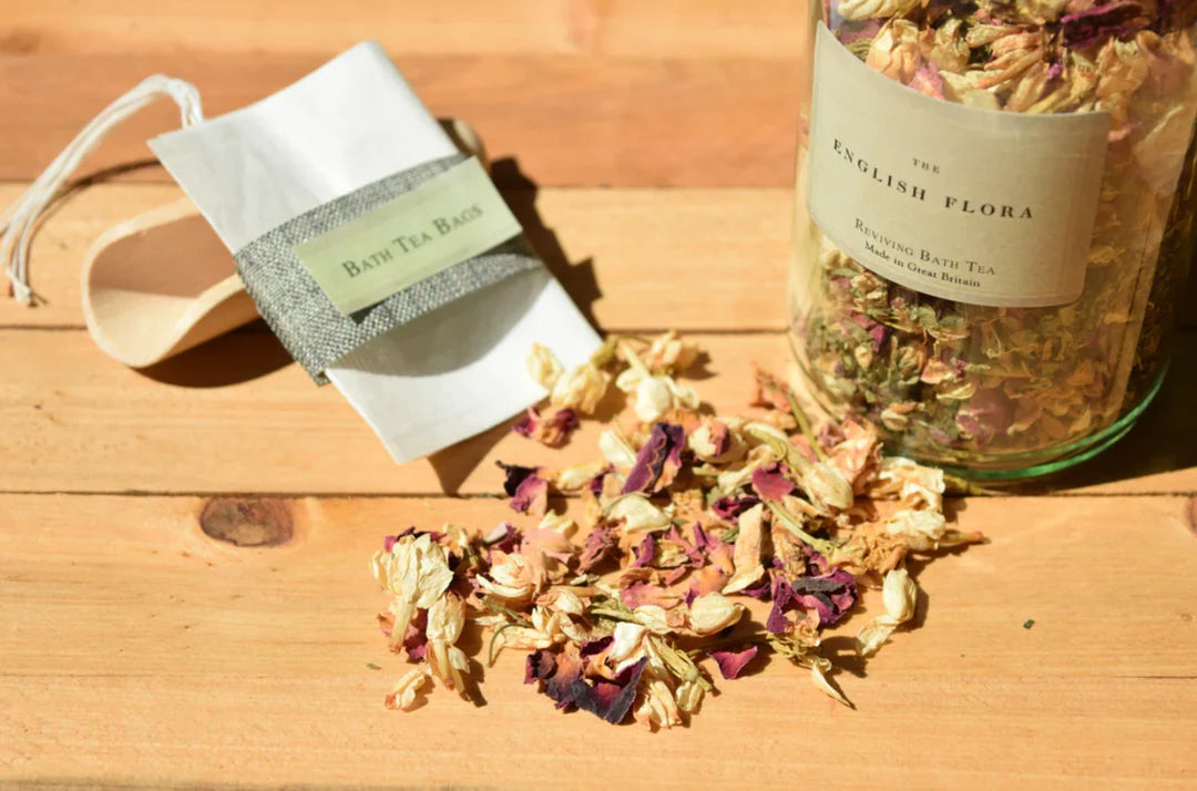Bath tea bags and jar of reviving bath tea in a glass jar labeled 'English Flora' and dried flowers on a wooden surface.
