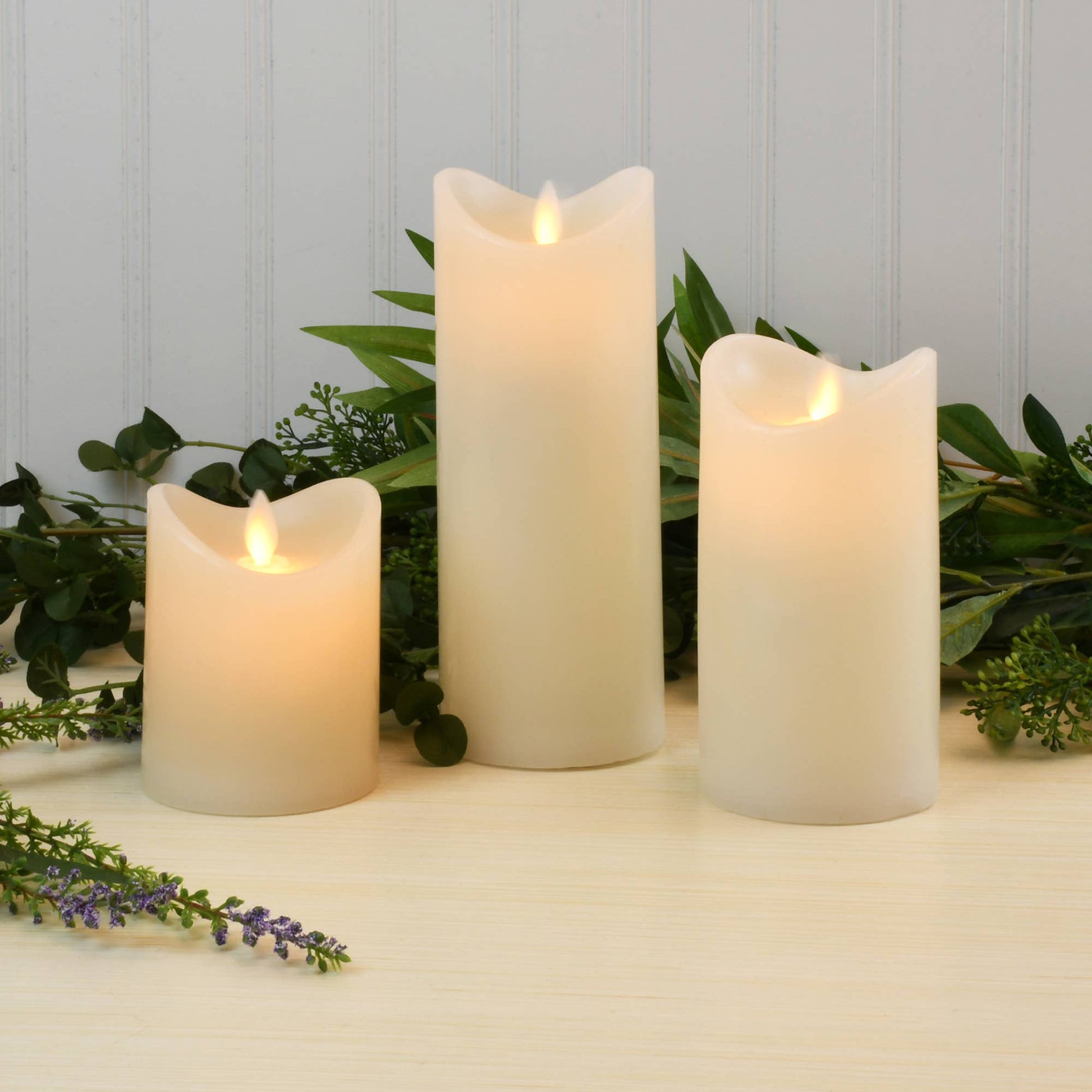 Three white flameless battery operated candles with greenery and lavender on a light surface and light blue background