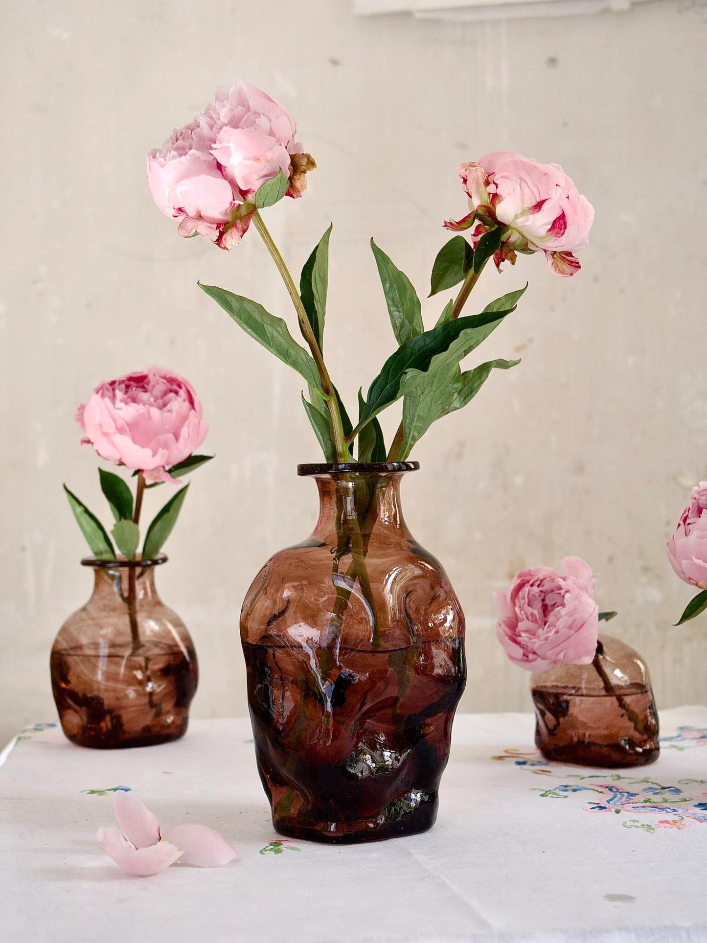 Three brown handmade glass vases with pink flowers on a light surface.