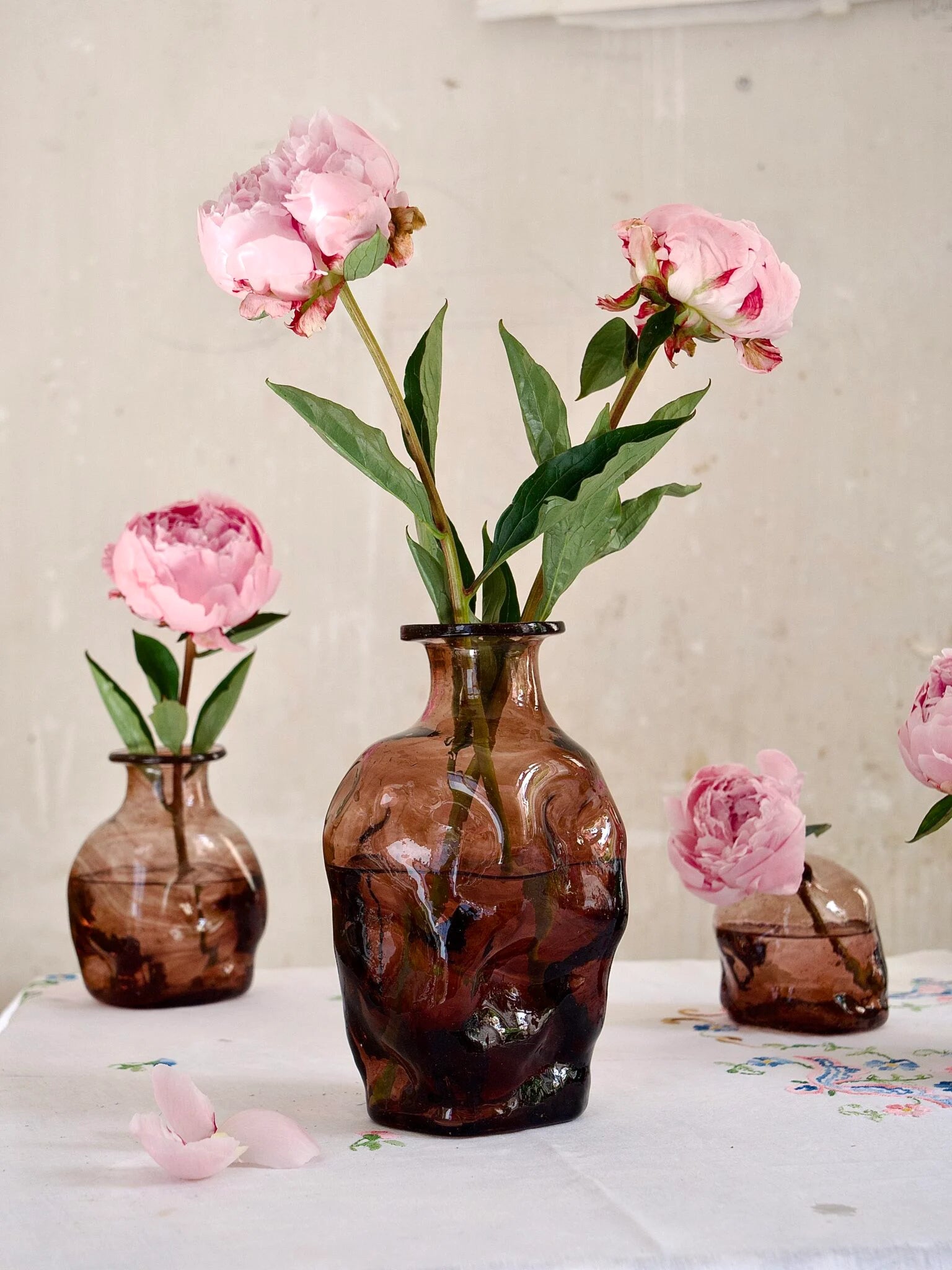 Three brown handmade glass vases with pink flowers on a light surface.