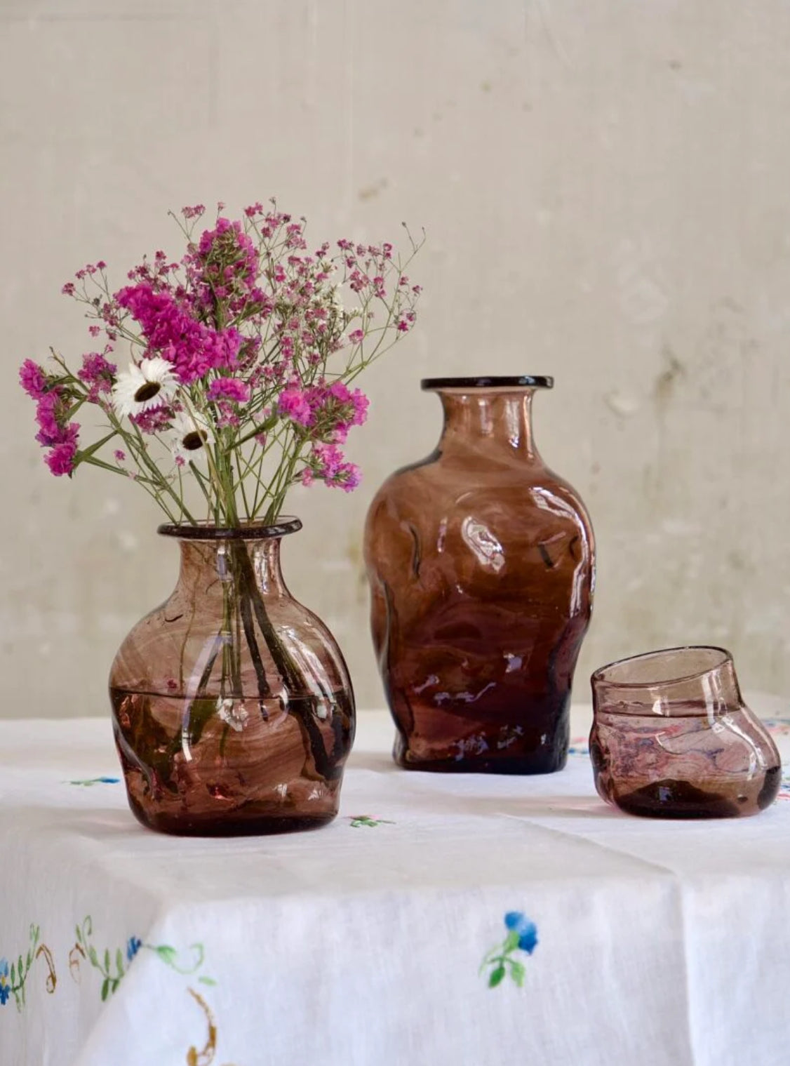 Two brown glass vases or carafes with a glass on a table with a floral arrangement.