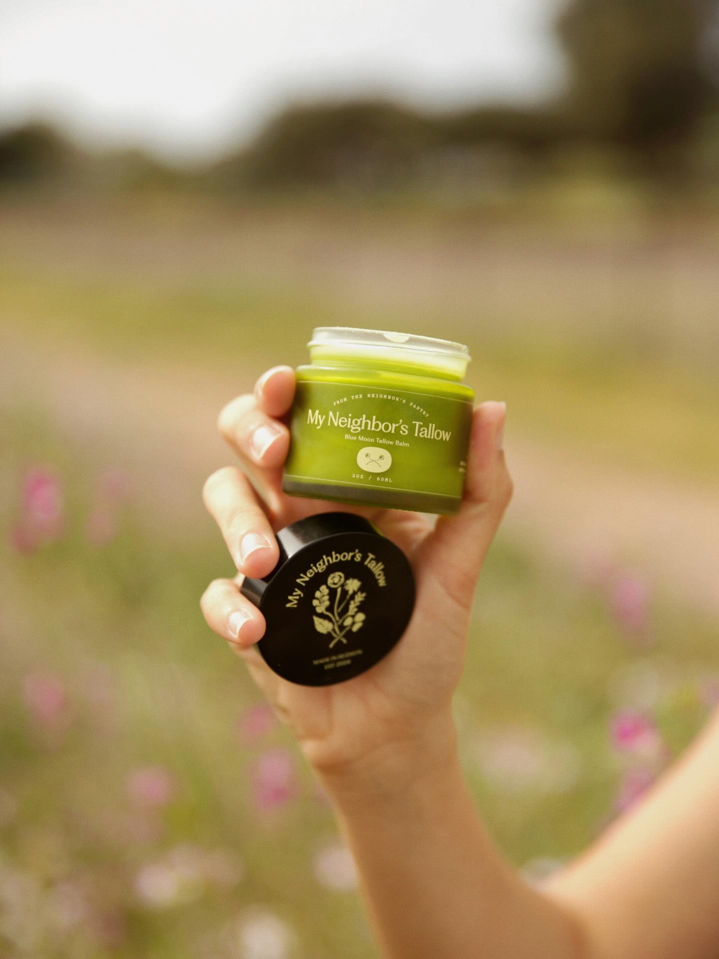 Hand holding a green jar with 'My Neighbor's Tallow' branding against a blurred natural background.