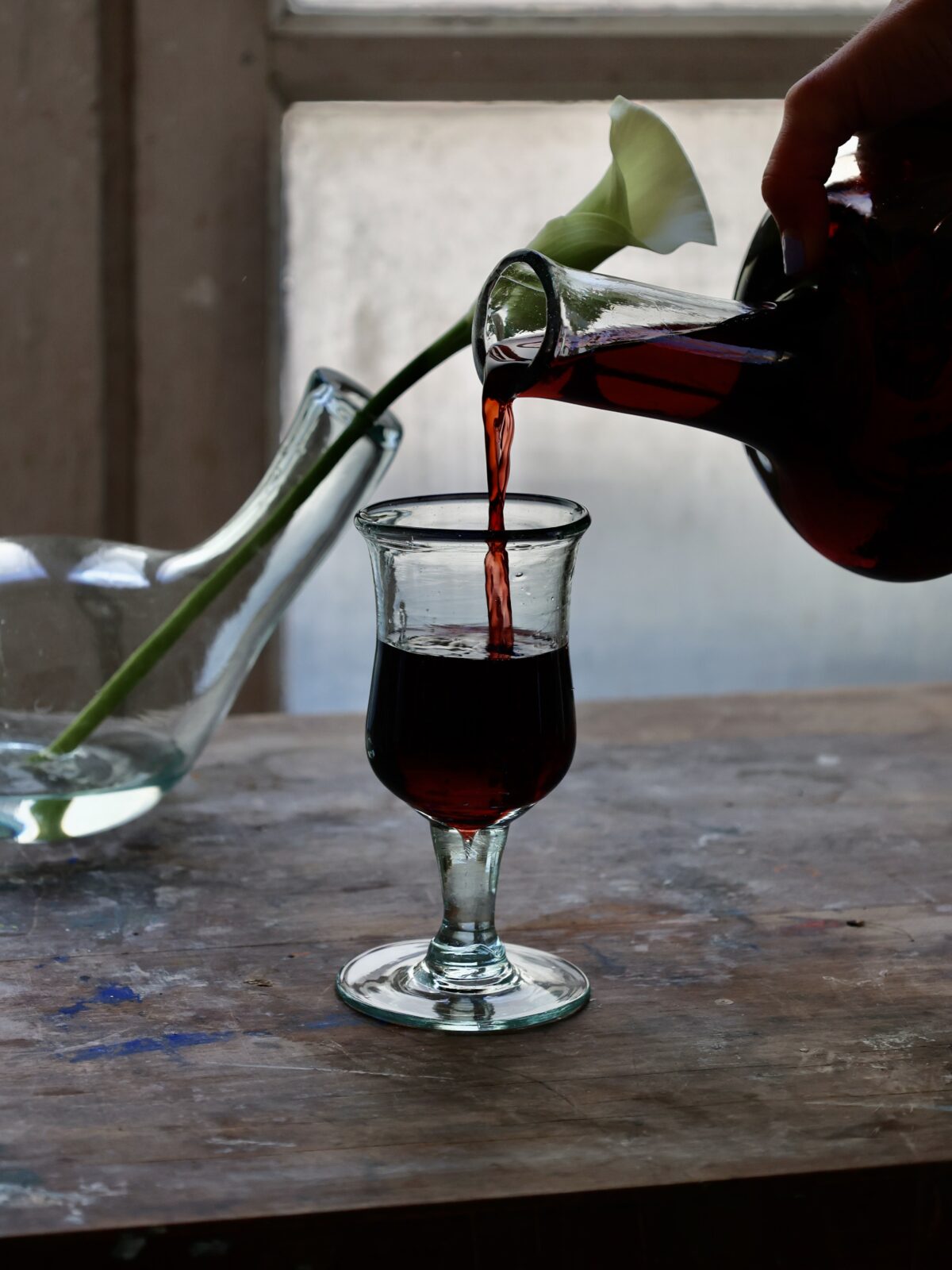 Pouring red wine from a bottle into a recycled glass wine glass crafted by artisans, with a white flower on a wooden surface.