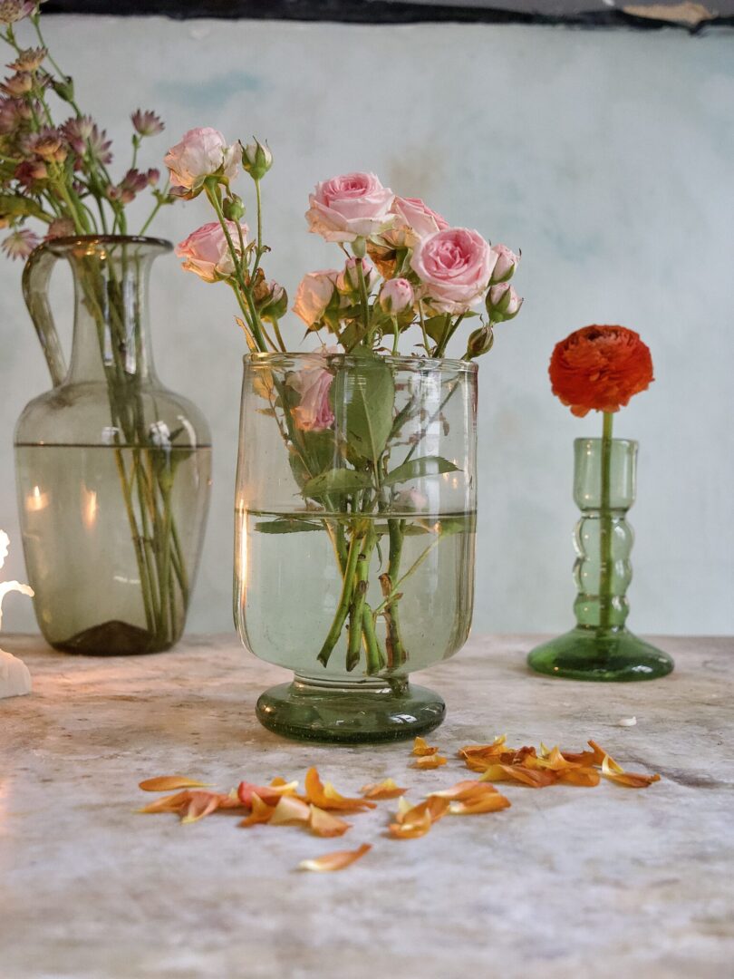 Three mouth-blown green glass vases with flowers on a wooden surface