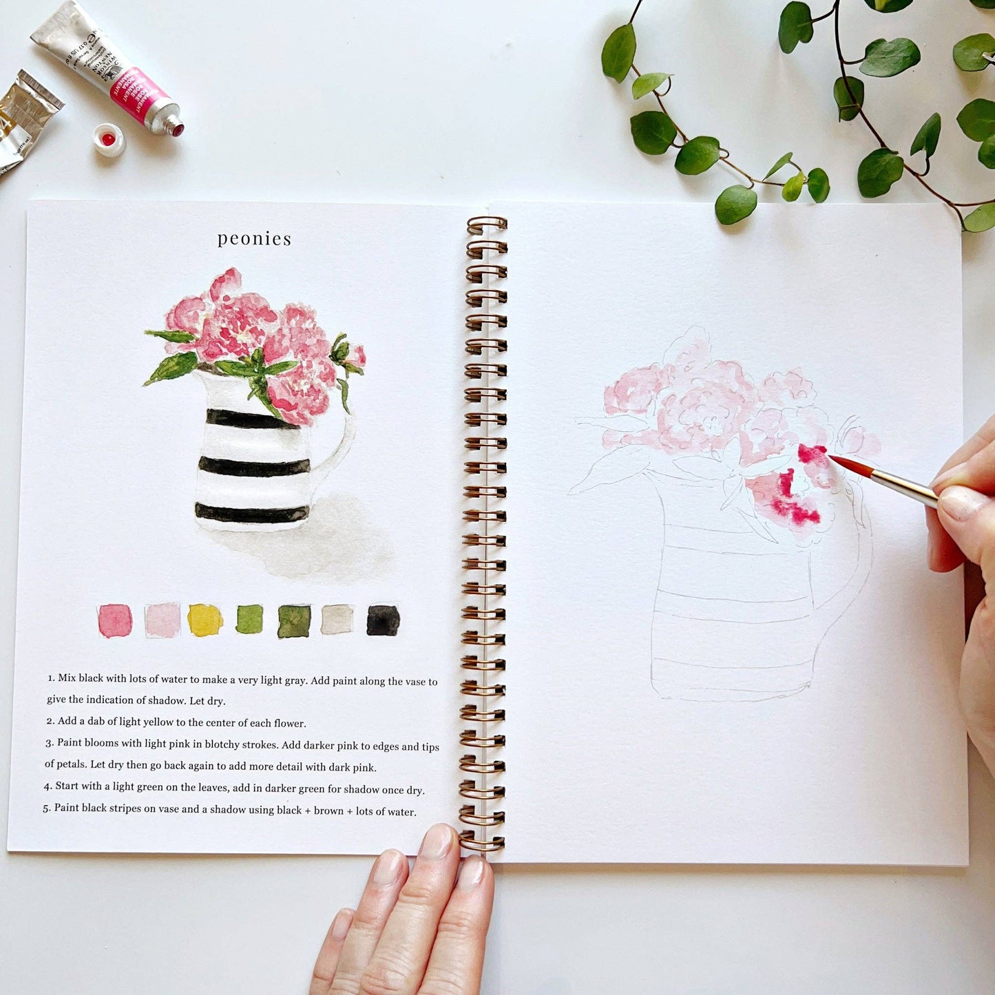 Open watercolor workbook with art lesson on flowers, hand holding paintbrush, and greenery.