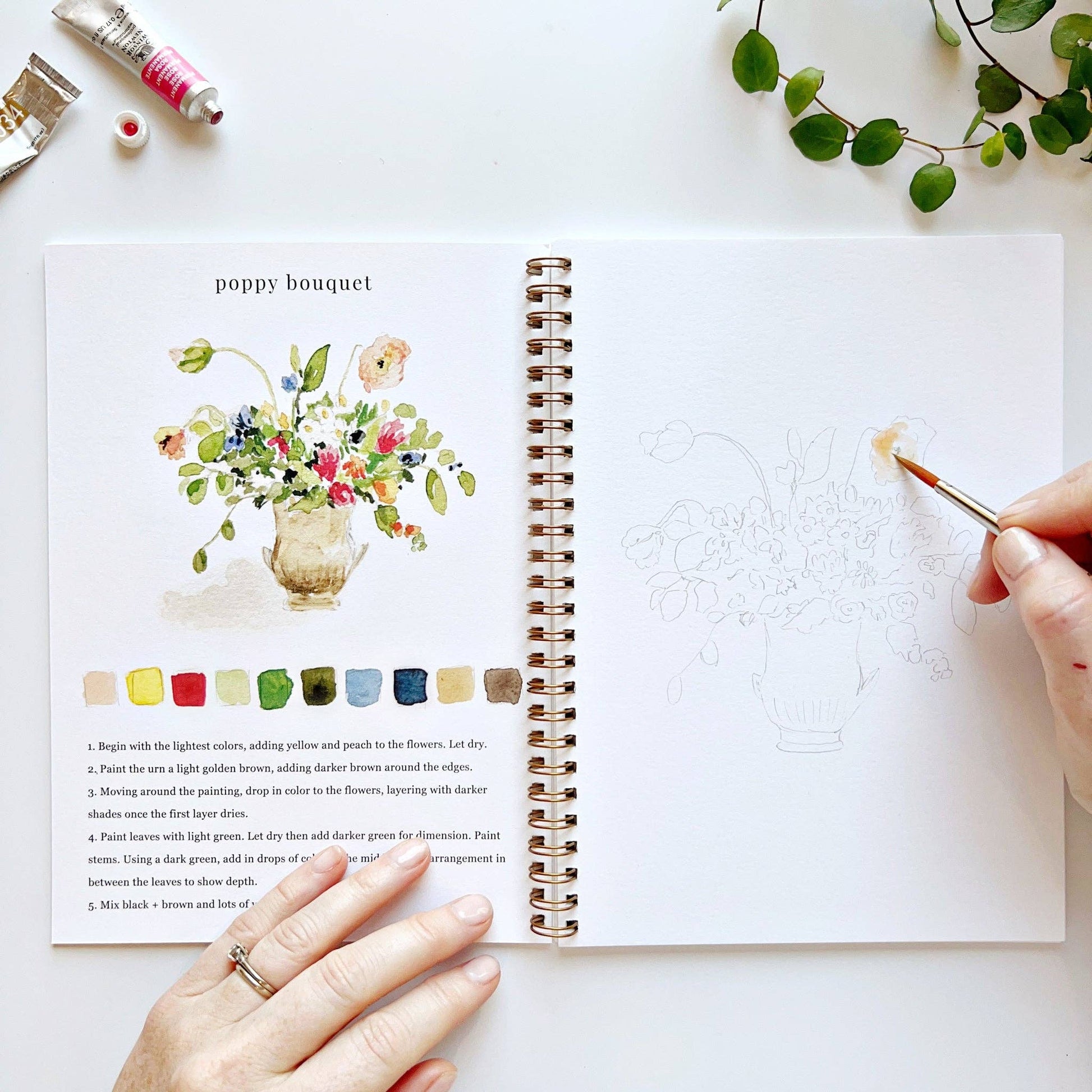 Artistic book with a painting of a poppy bouquet, color swatches and step-by-step instructions, with a hand holding a paintbrush.