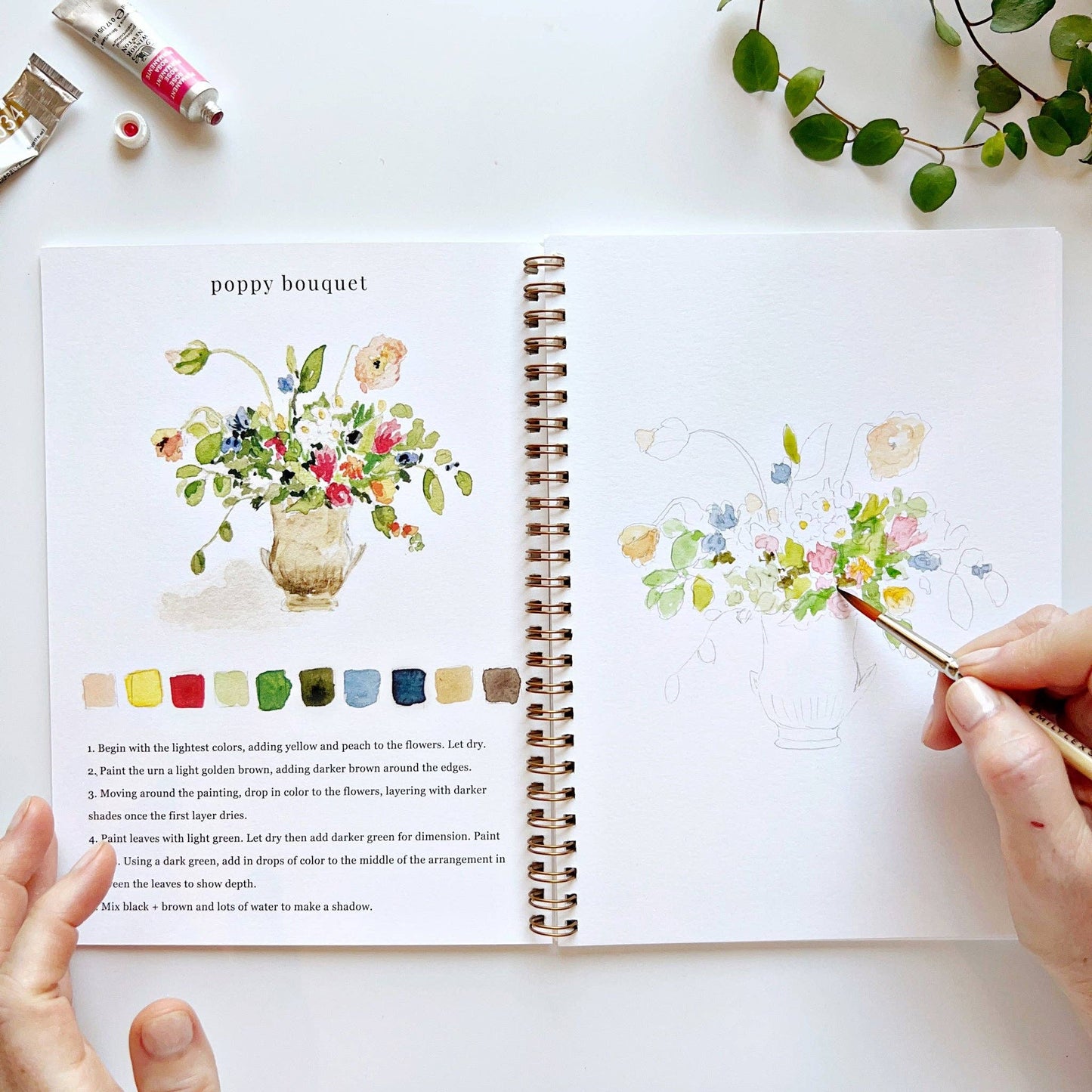 Open watercolor workbook with floral illustrations and color swatches, being used as a reference for painting.