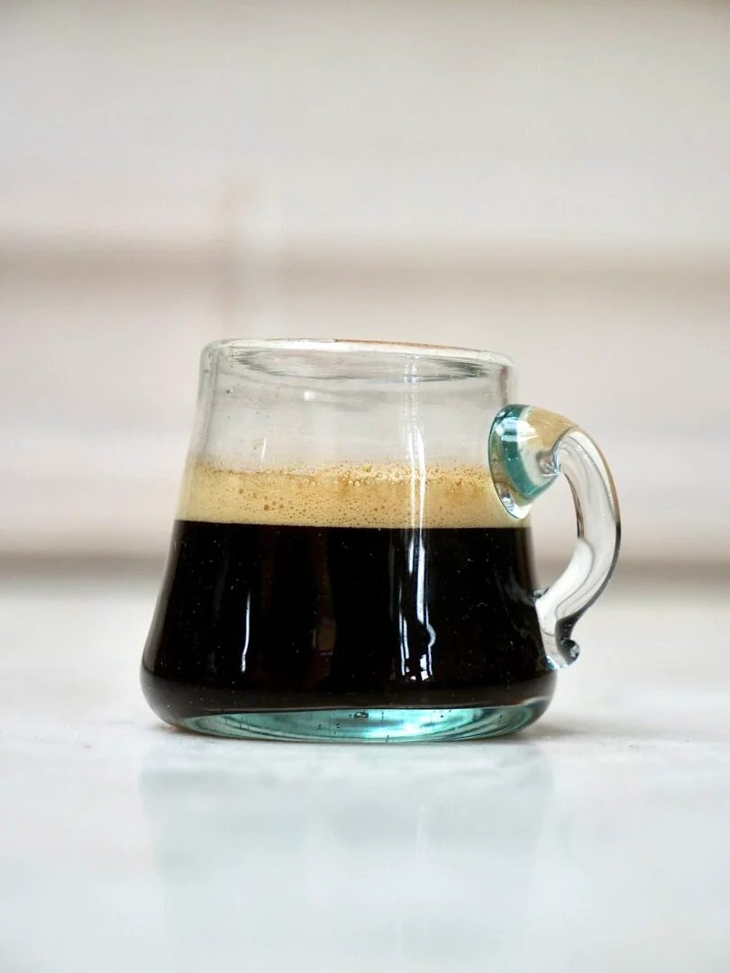 Clear mouth-blown glass mug with a handle filled with coffee on a light background