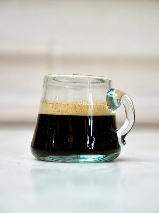 Clear mouth-blown glass mug with a handle filled with coffee on a light background