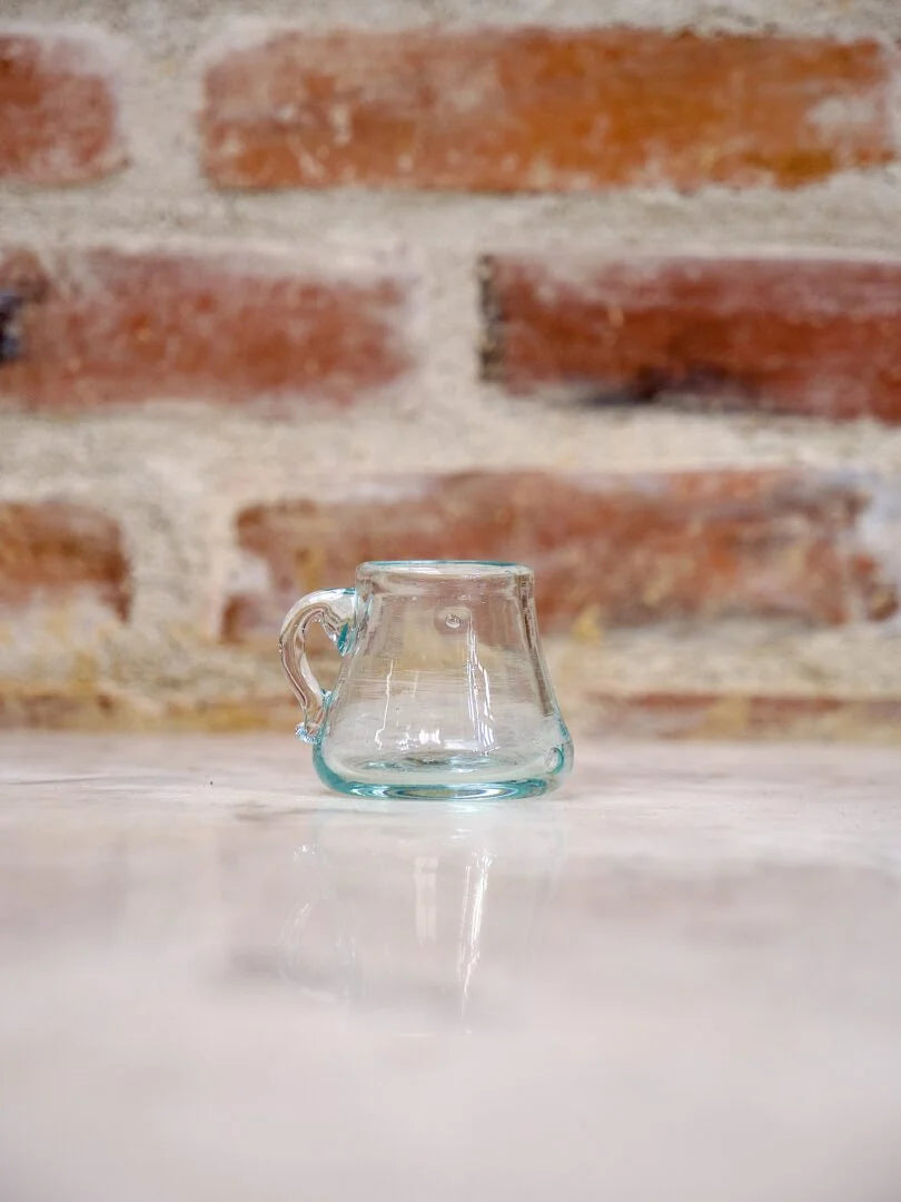 Clear hand-blown glass mug on a white surface with a brick wall background