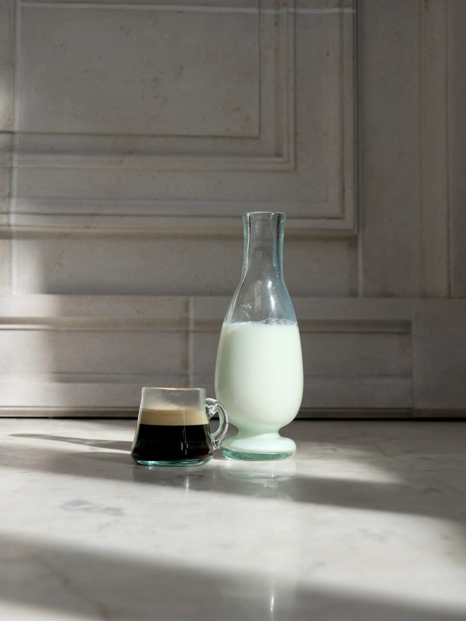 Glass carafe filled with milk next to a glass cup of coffee on a marble surface. They both are artisanal and mouth-blown.