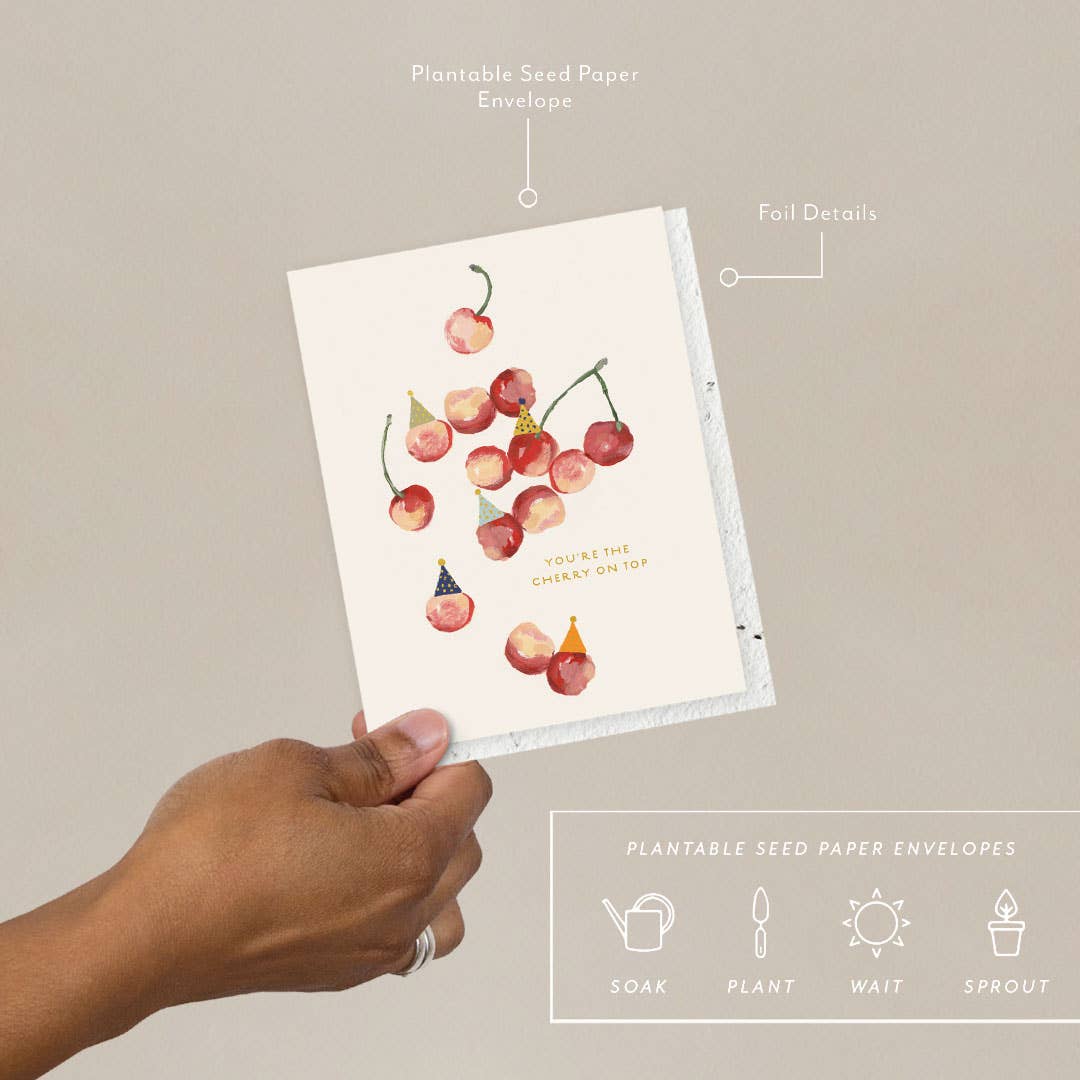 A hand holding a greeting card with an illustration of cherries wearing party hats, with a text that reads 'You're the cherry on top', accompanied by a plantable seed paper envelope with eco-friendly packaging details.
