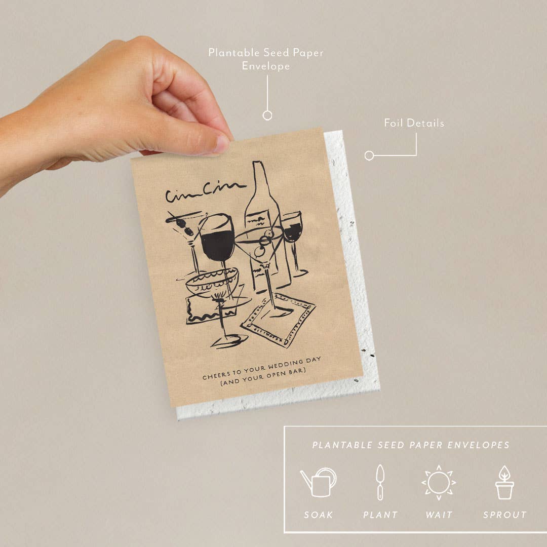 A hand holding a beige-colored greeting card with a plantable seed paper envelope, featuring foil details and a wedding-themed design with cocktails.
