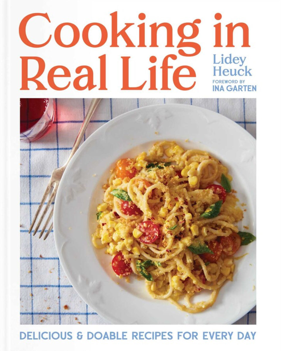 Book cover of 'Cooking in Real Life' by Lidey Heuck, with a foreword from Ina Garten. Delicious and doable recipes for every day, picturing a plate of pasta and vegetables on a checkered tablecloth.
