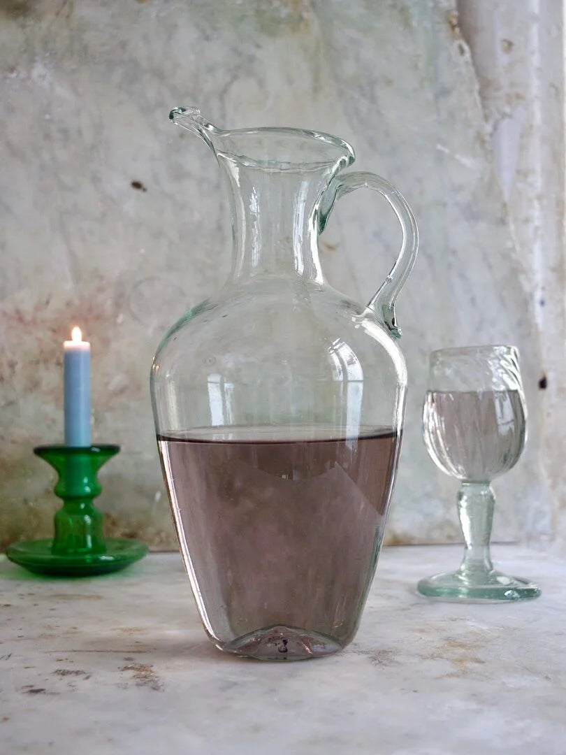 Clear glass pitcher with liquid on a rustic surface with a candle and glass in the background.