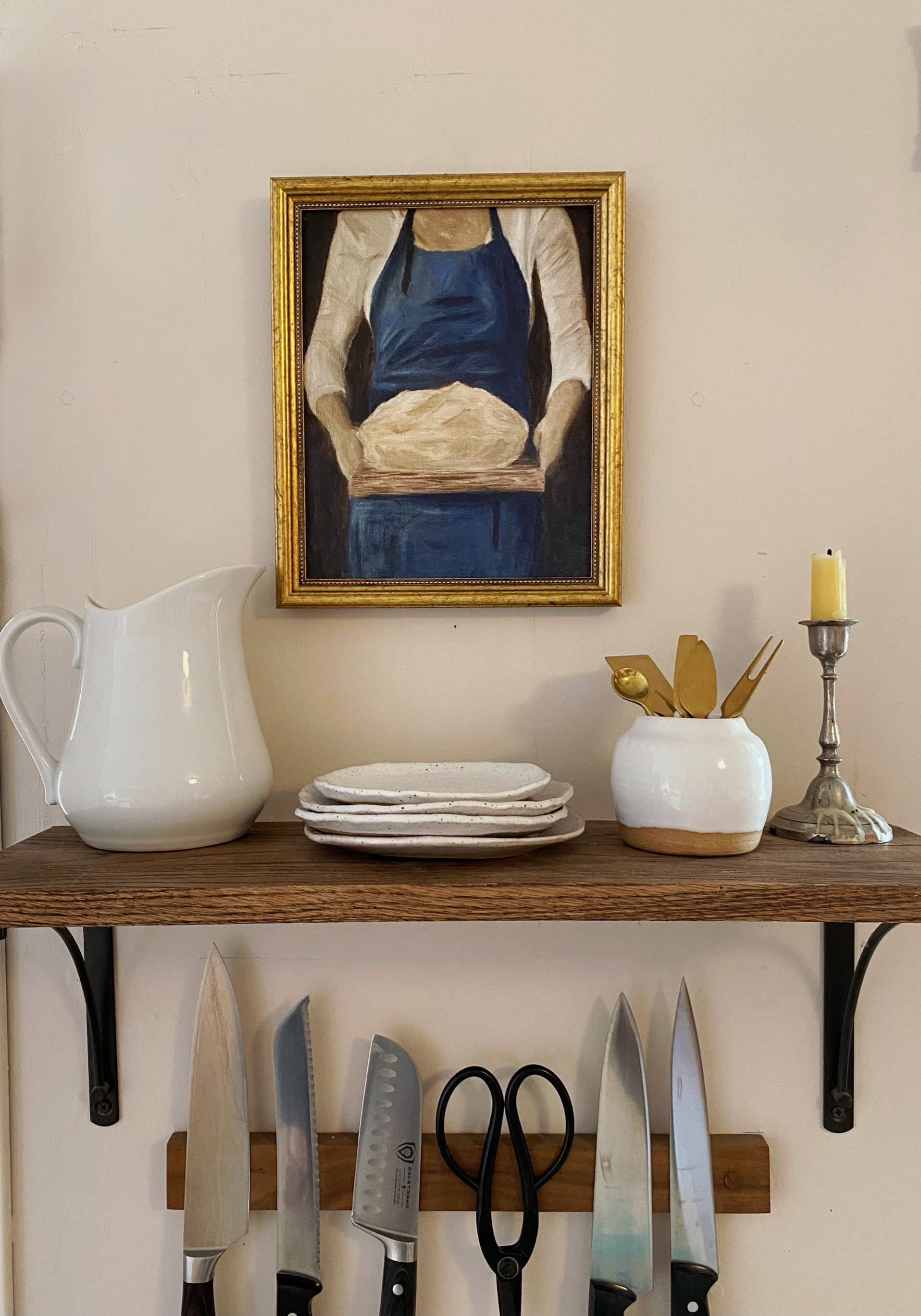 French-inspired art print of a woman holding sourdough bread, here hanged on a kitchen wall, with plates and cutlery beneath it.