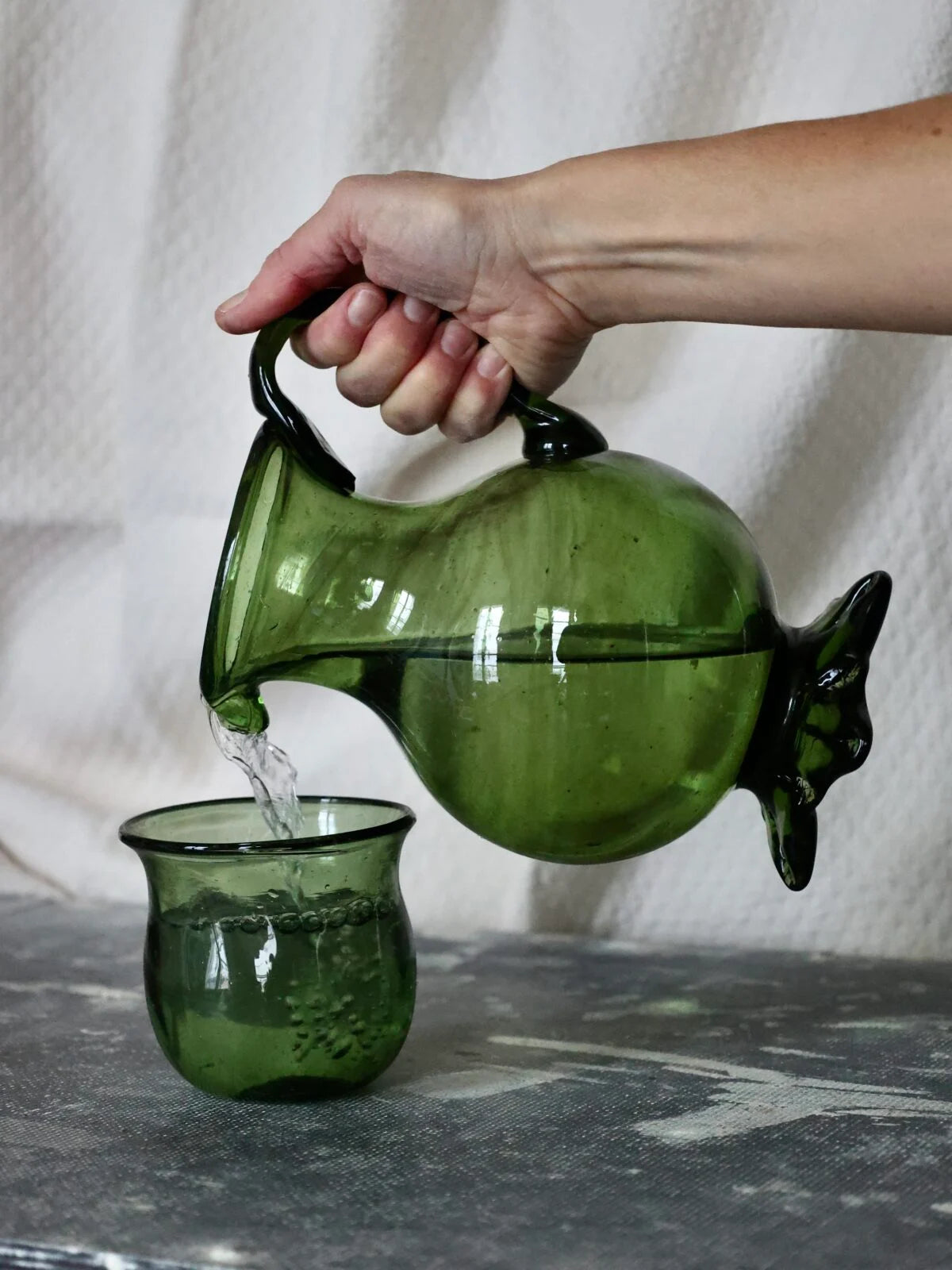 Green glass carafe or pitcher being used to pour water into a matching glass cup on a stone surface.