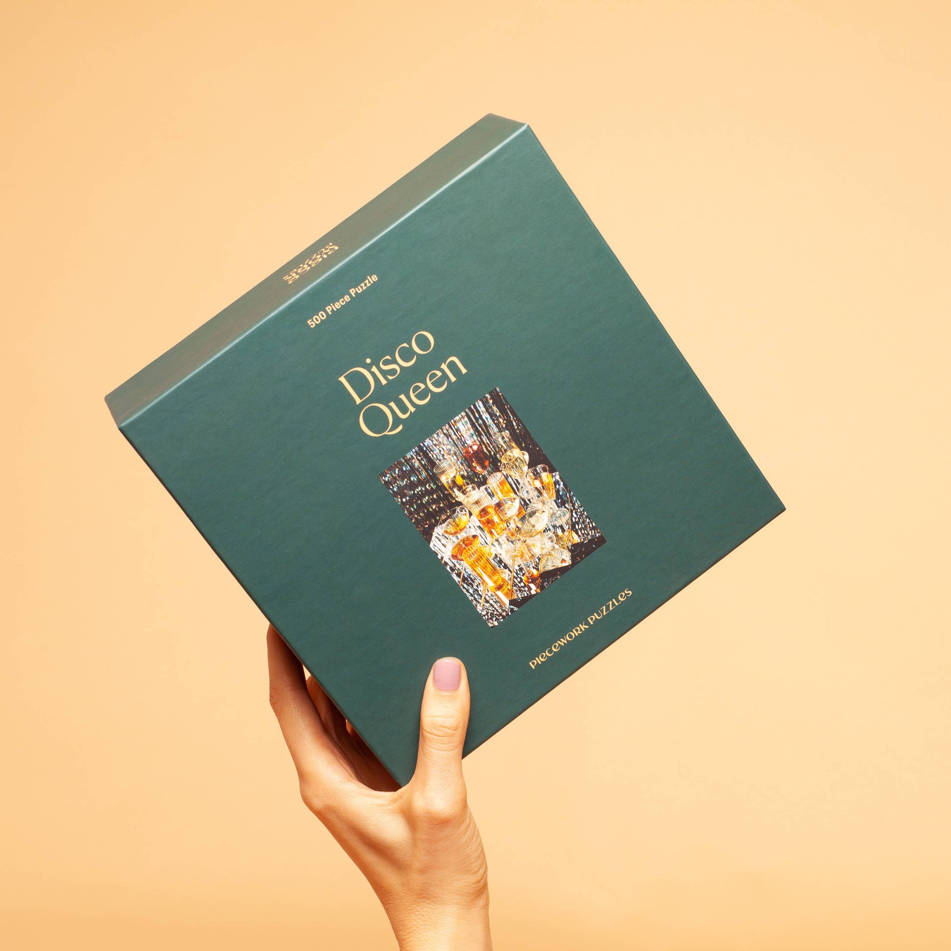 Disco Queen 500-piece puzzle box with colorful artwork depicting glasses and cocktails from Piecework Puzzles, held in a hand.