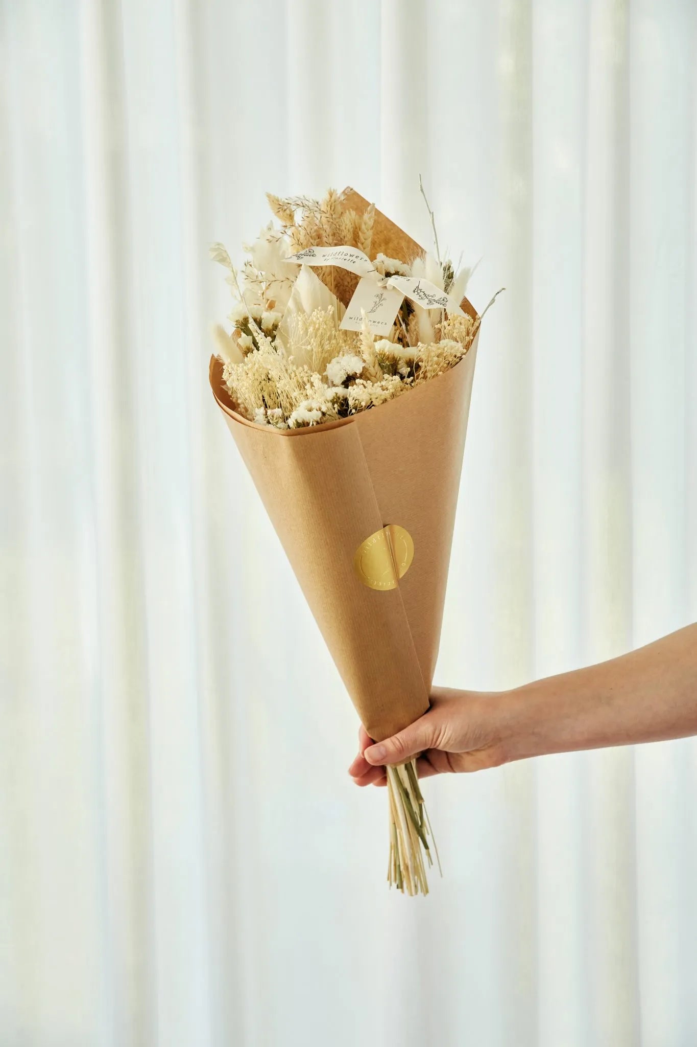 Bouquet of natural dried flowers wrapped in brown paper with gold dots, held against a white curtain background.