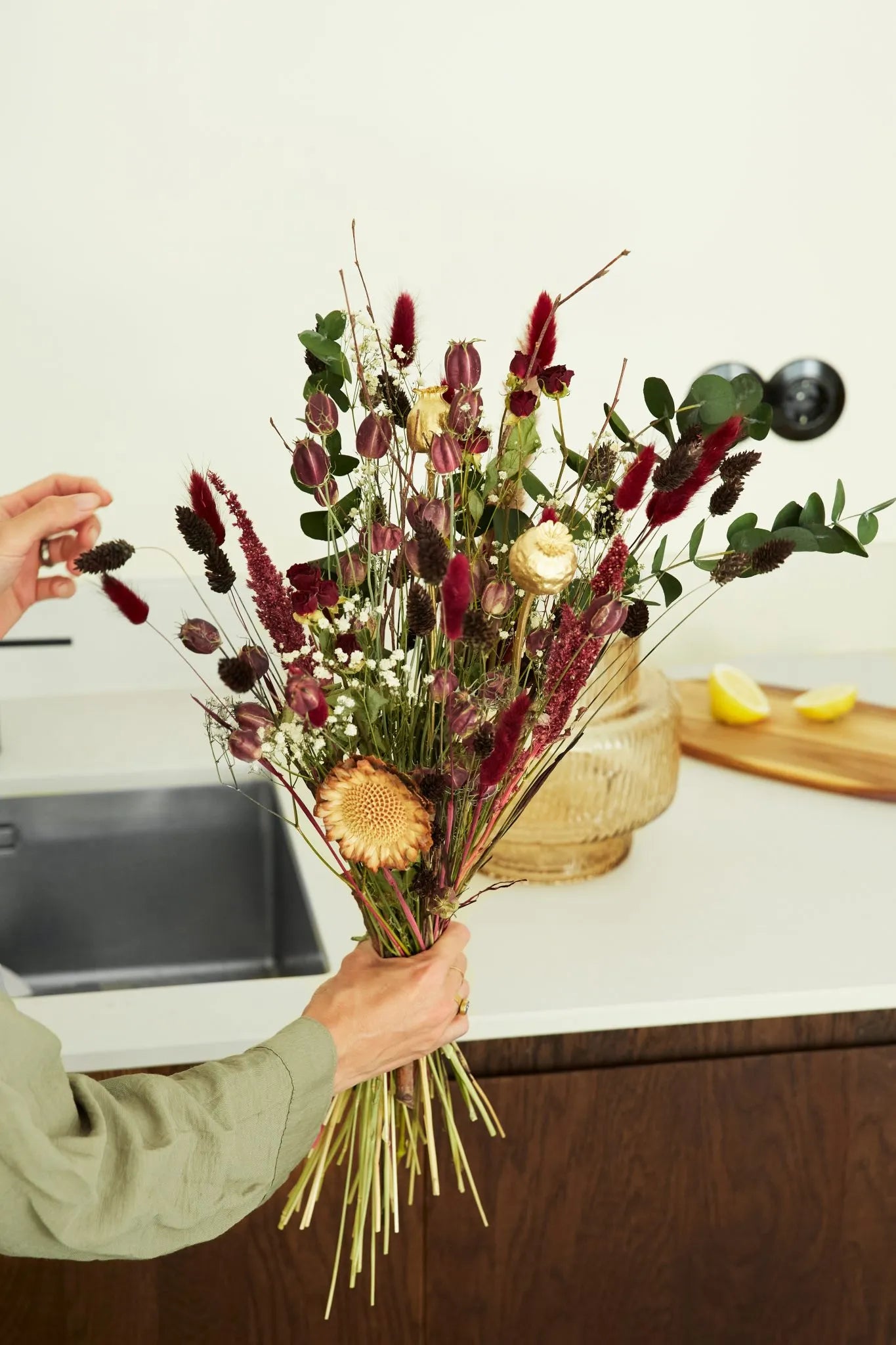 Person holding a bouquet of natural dried flowers in a kitchen. Tones of colors are red and green.