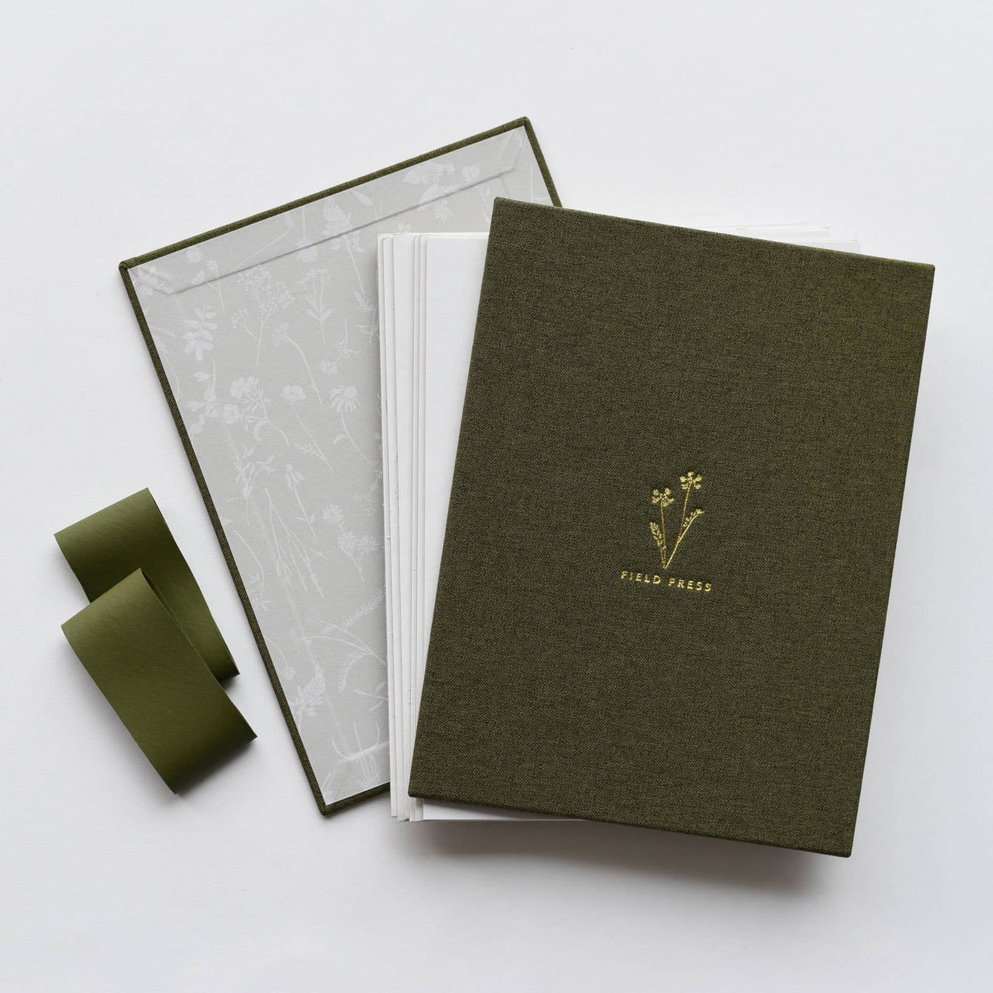 A green linen-covered field flower press with gold foil details, open with seven reusable recycled pressing cards and two heavyweight bands by June & December.