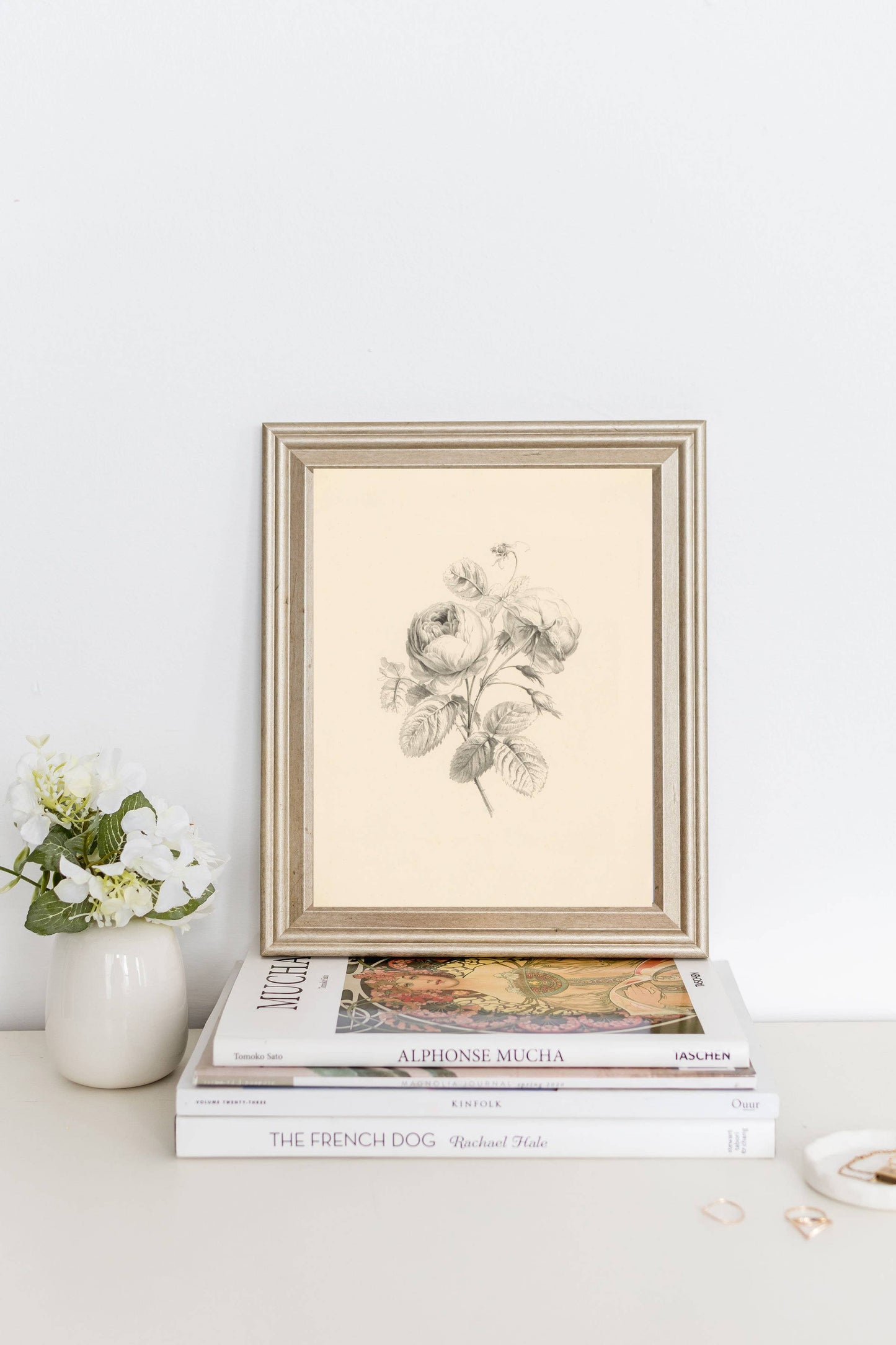 Vintage botanical wall art reproduction depicting flowers, displayed on a stack of books with some real flowers nearby, on a white background.