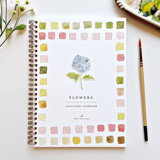 A watercolor workbook titled 'FLOWERS' with a watercolor illustration of a blue hydrangea on the cover, surrounded by watercolor color swatches. A watercolor brush is lying next to the book.