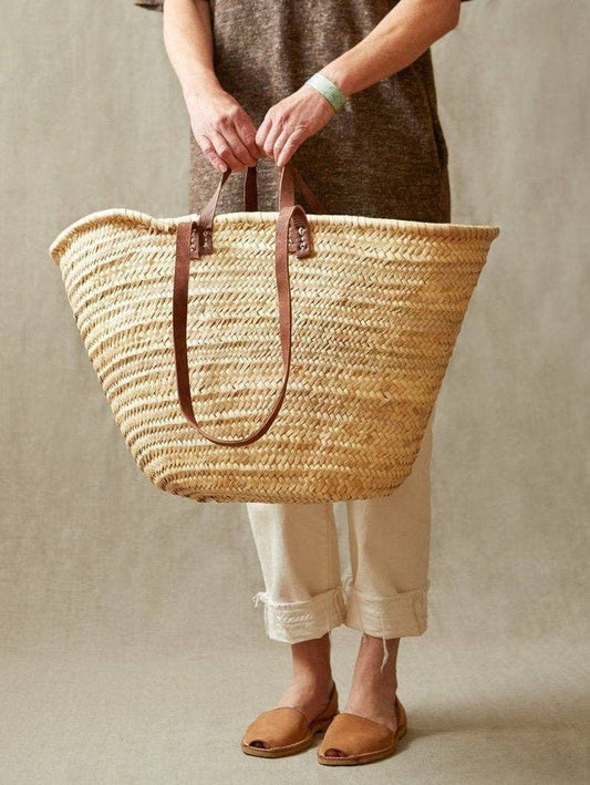 Person holding a woven straw market basket with leather handles against a neutral background