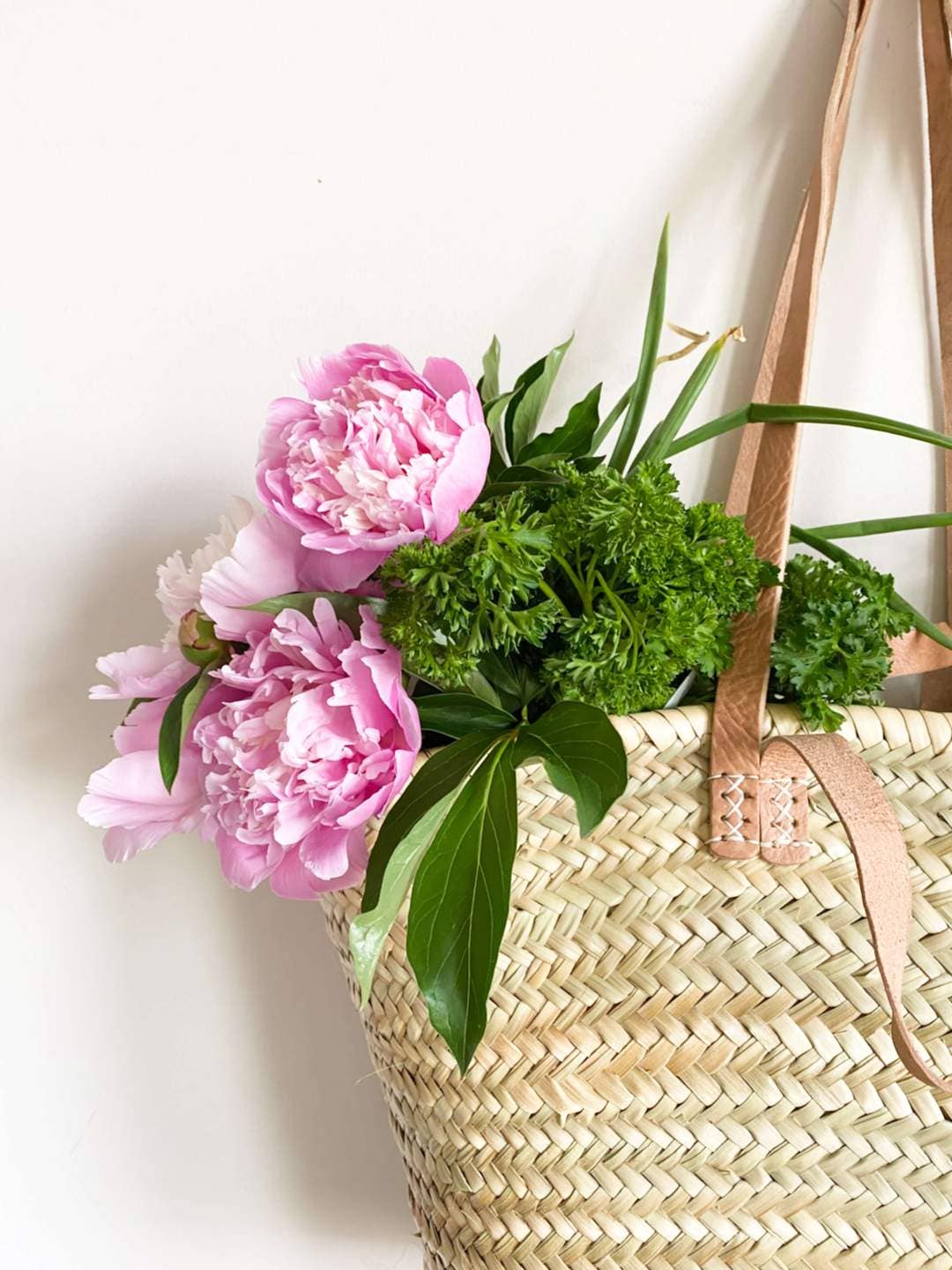 Handwoven Moroccan straw basket with leather handles with pink flowers and greenery on a white background
