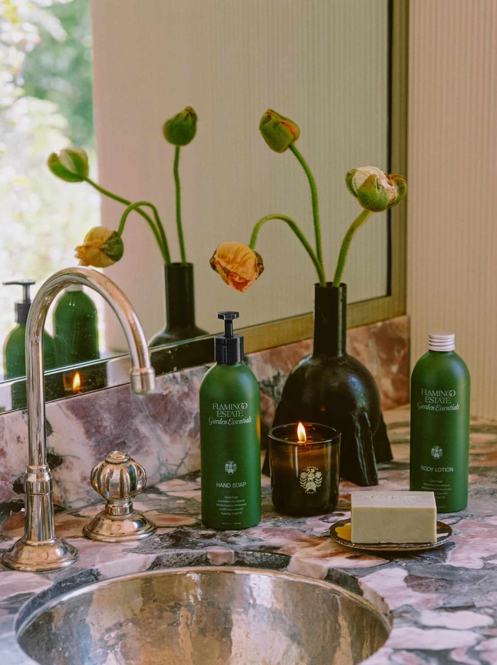 Bathroom setting with green Flamingo Estate Hand Soap bottles, a candle, and flowers on a marble countertop.