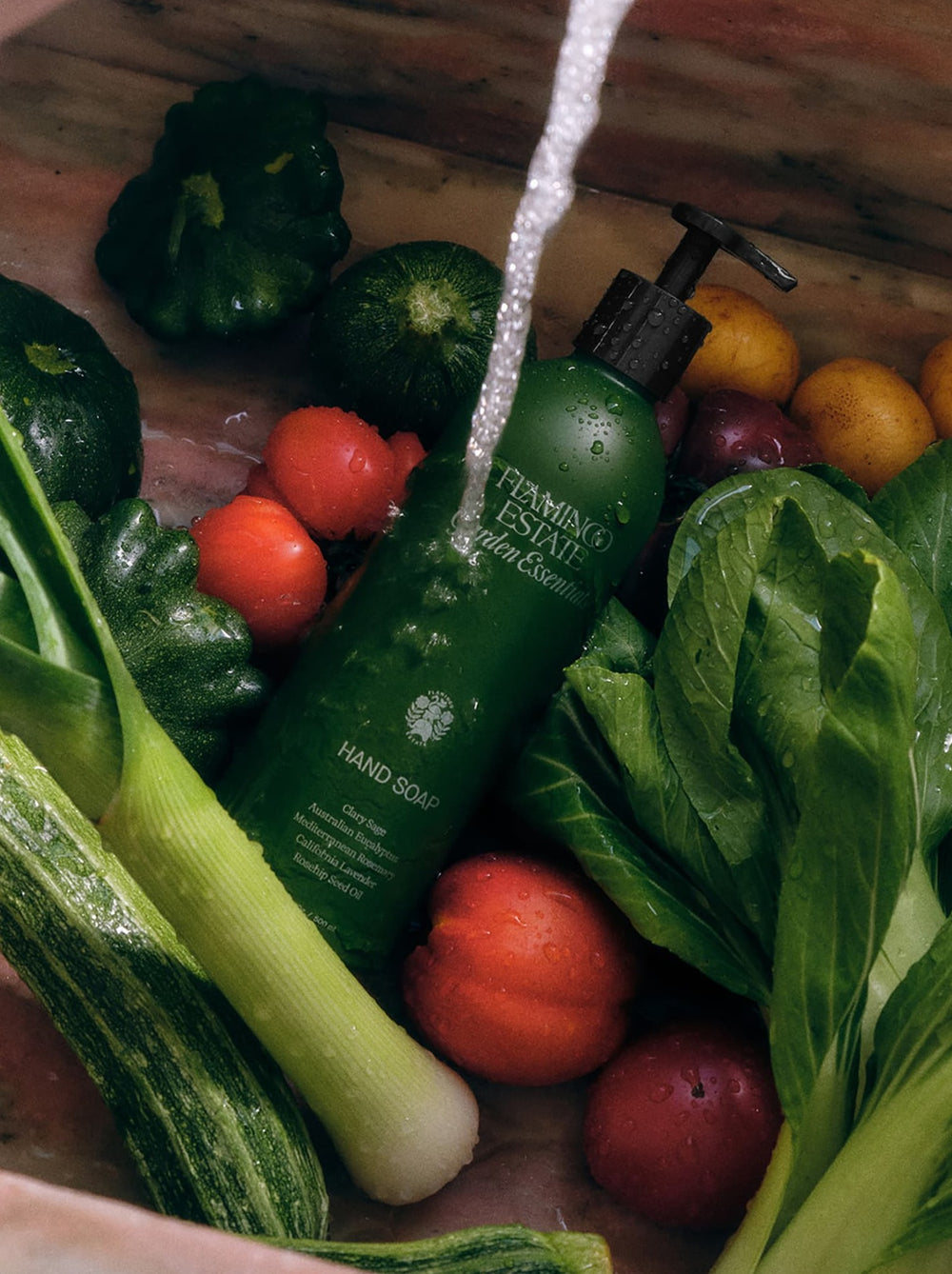Green bottle of Flamingo Estate hand soap among vegetables on a wooden surface