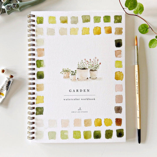 A watercolor workbook titled 'GARDEN' with plant illustrations and a variety of watercolor swatches on the cover, alongside a paintbrush and watercolor paints.