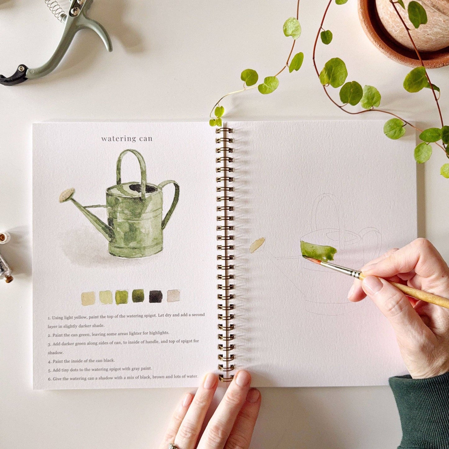 A garden watercolor workbook with step-by-step garden sketches, here on the watering can page, with someone painting with a brush