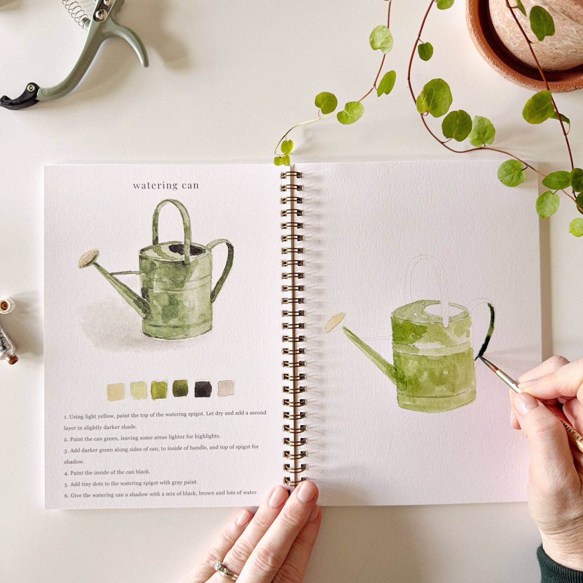 A garden watercolor workbook with step-by-step garden sketches, here on the watering can page, with someone painting with a brush, with a plant next to the workbook
